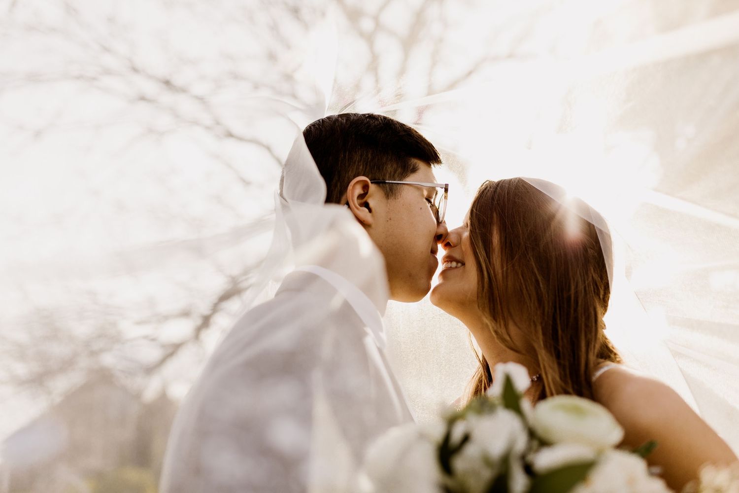 A bride and groom share a romantic kiss under a flowing veil with soft sunlight and bare trees.