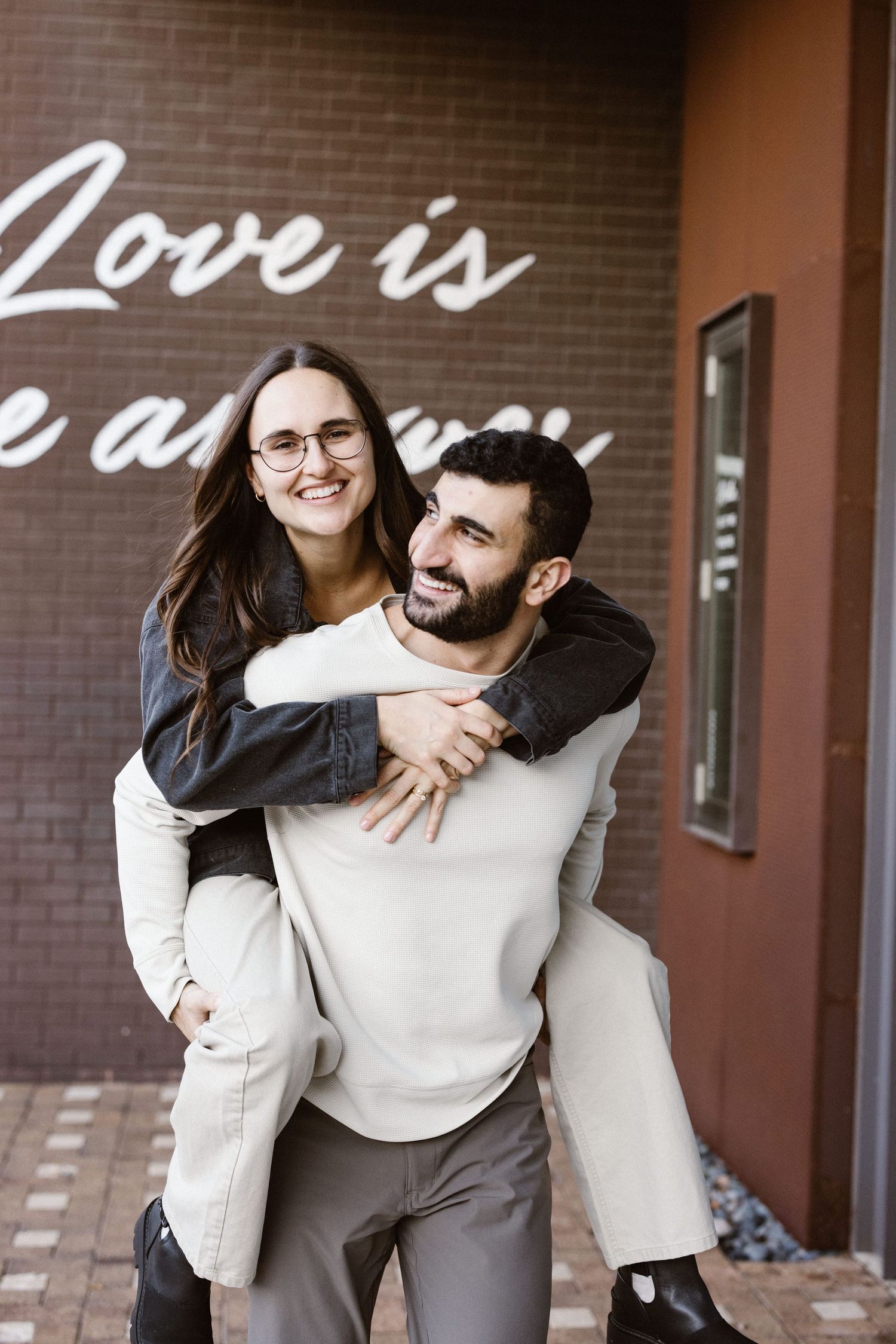 Woman piggyback riding man in front of a brick wall with Love Is sign.