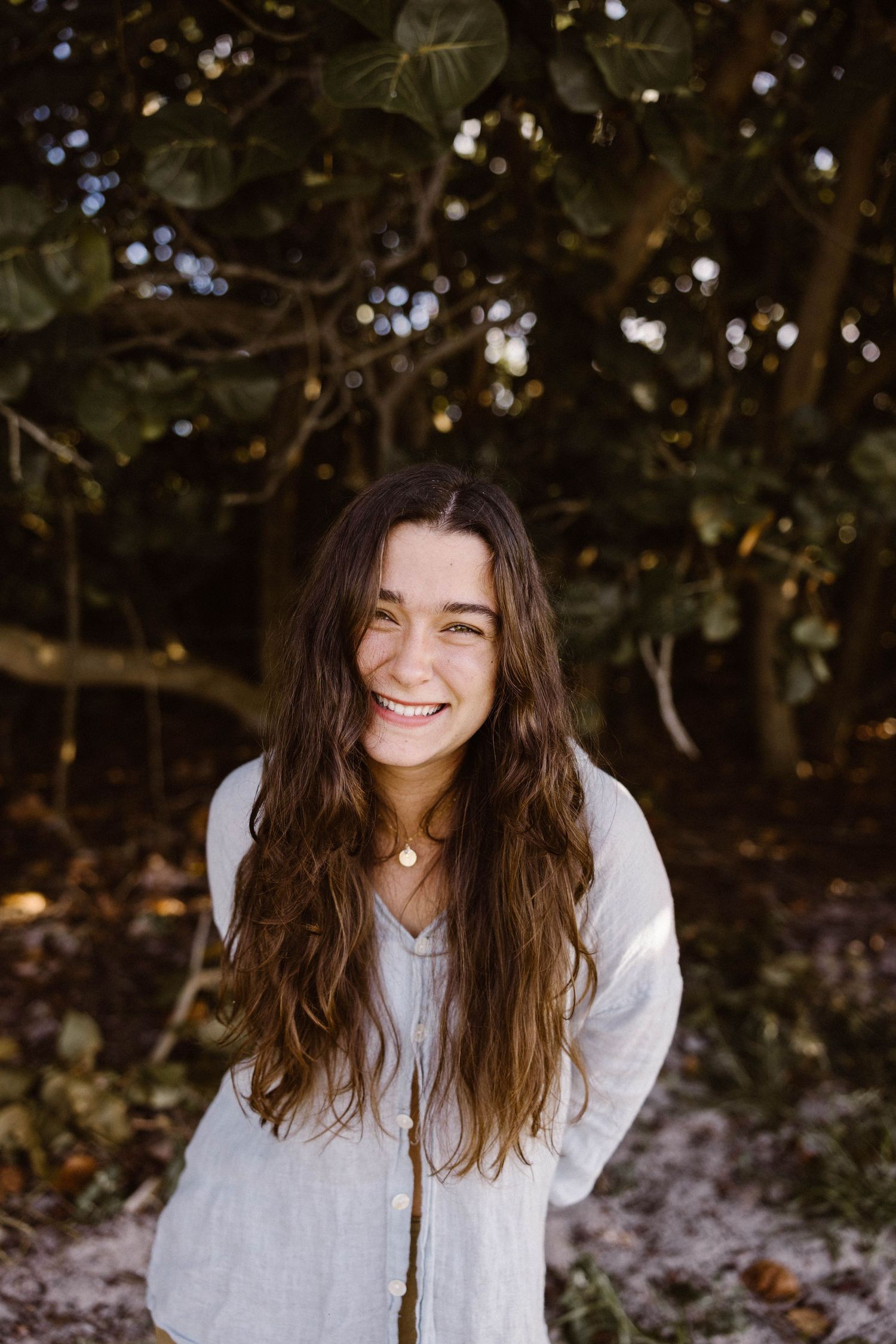 Smiling young woman with long brown wavy hair wearing a white linen shirt outdoors near trees.