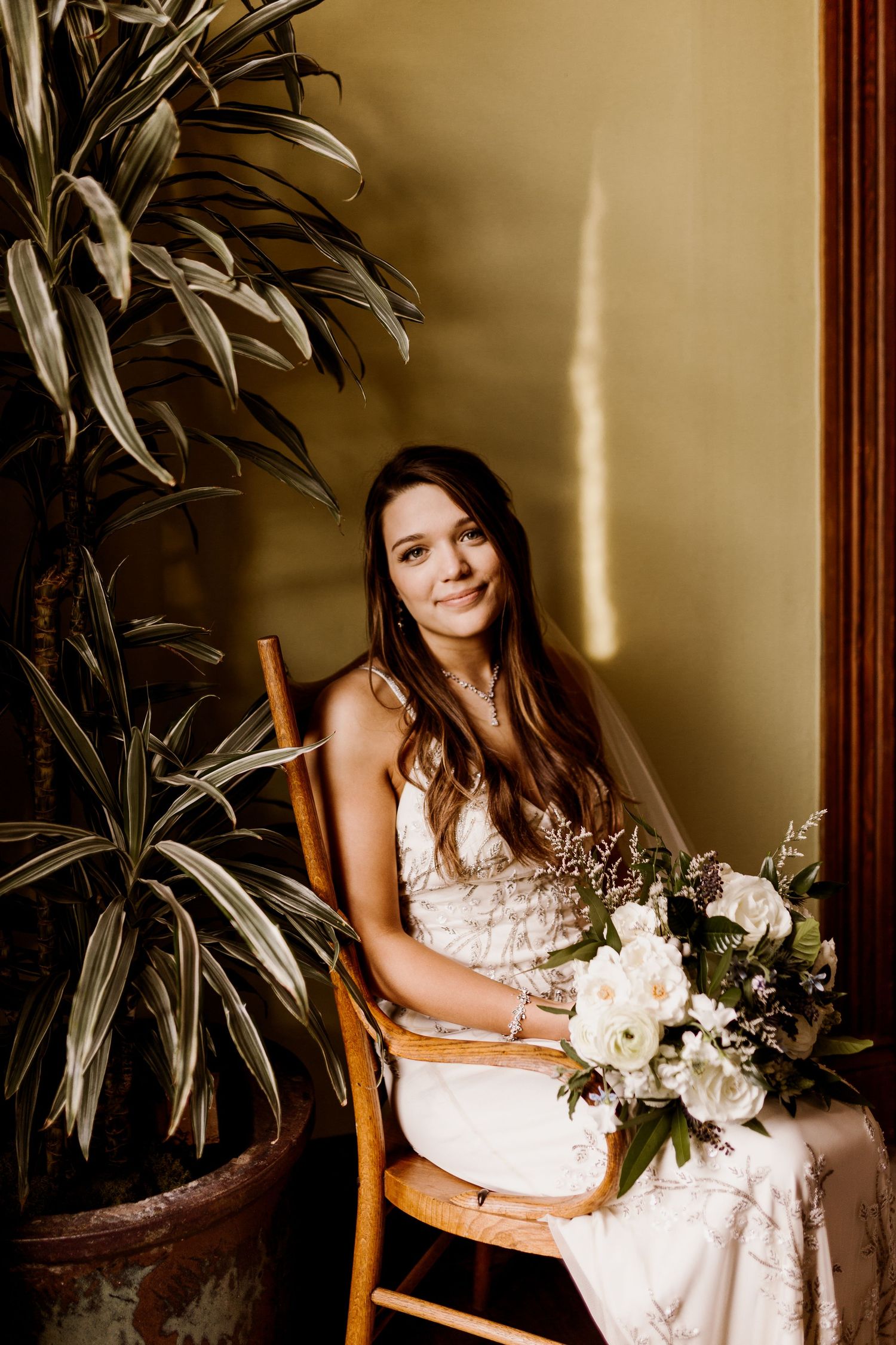 Smiling bride in lace wedding dress seated in wooden chair holding white bouquet beside tall houseplant.