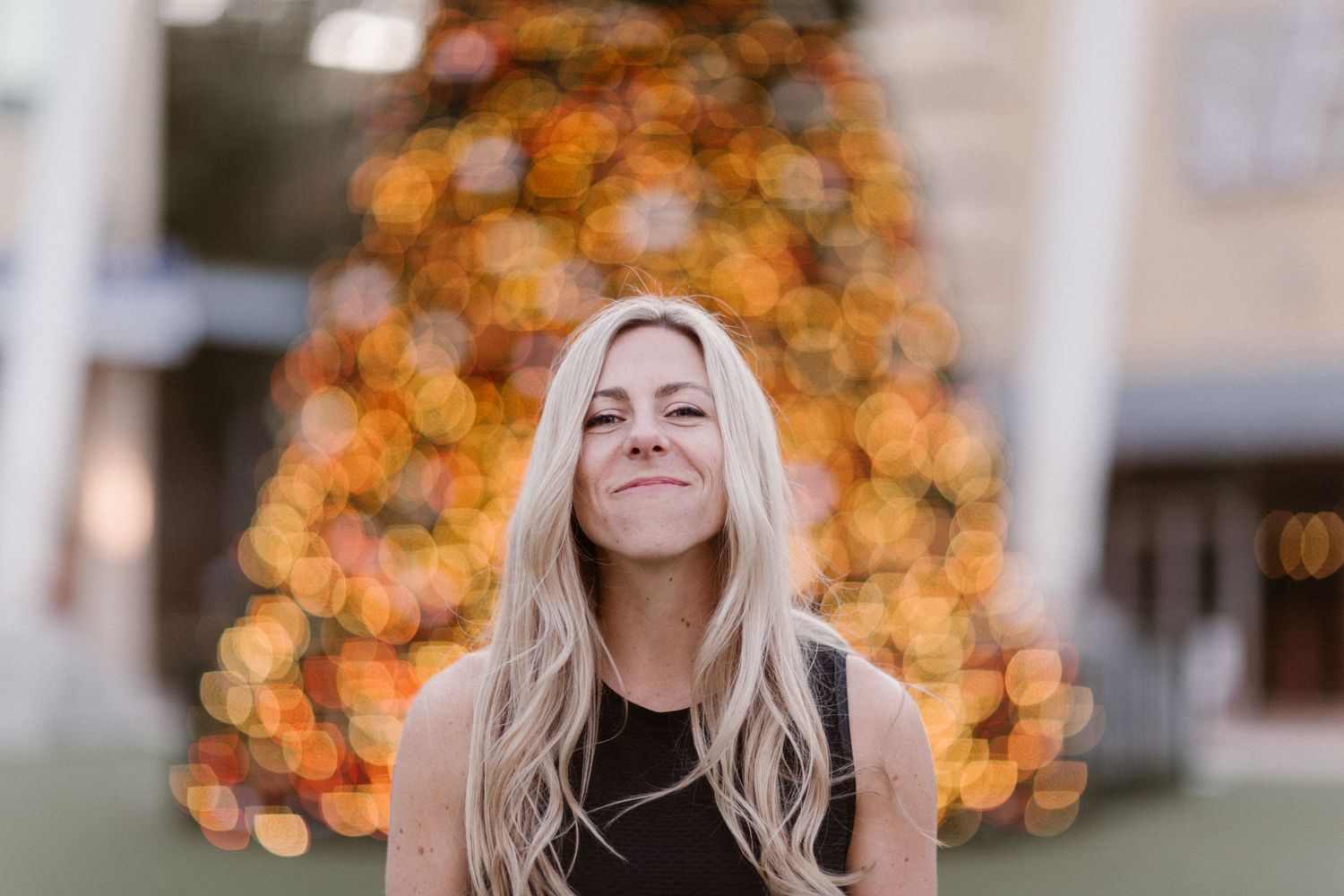 Smiling blonde woman in black top posing outdoors with bokeh Christmas tree lights in background.