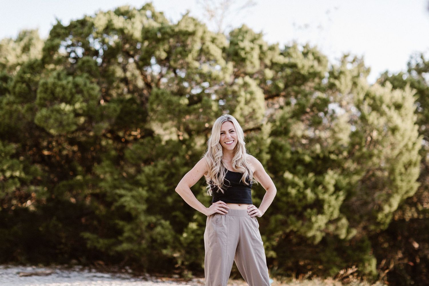 Smiling blonde woman in black top and tan wide-leg pants posing outdoors with trees in background.