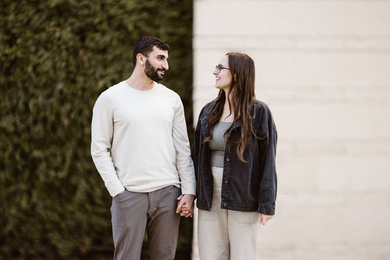 Smiling couple holding hands and gazing at each other while standing in front of a green hedge wall.