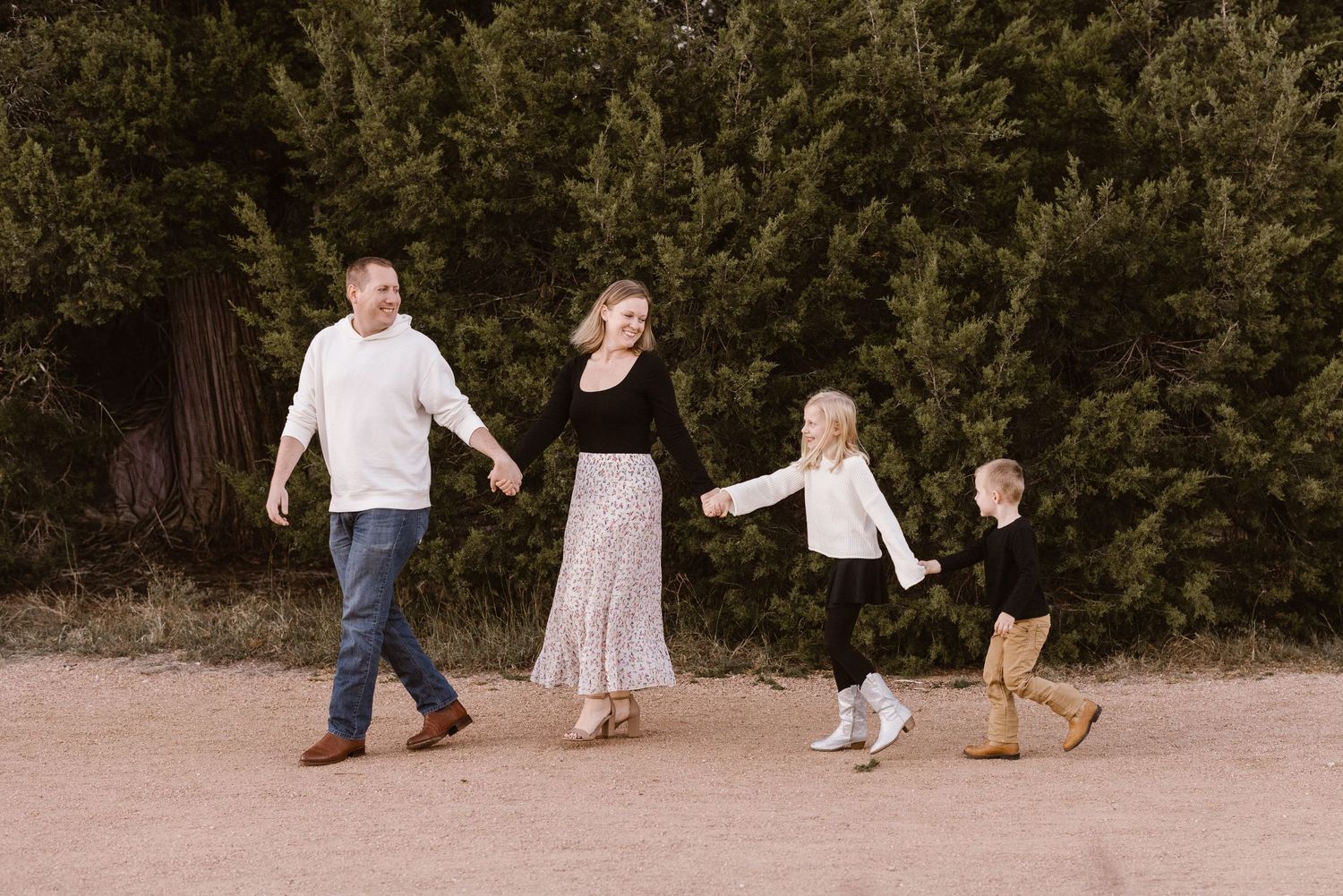 Joyful family of four holding hands and walking together near tall green trees.