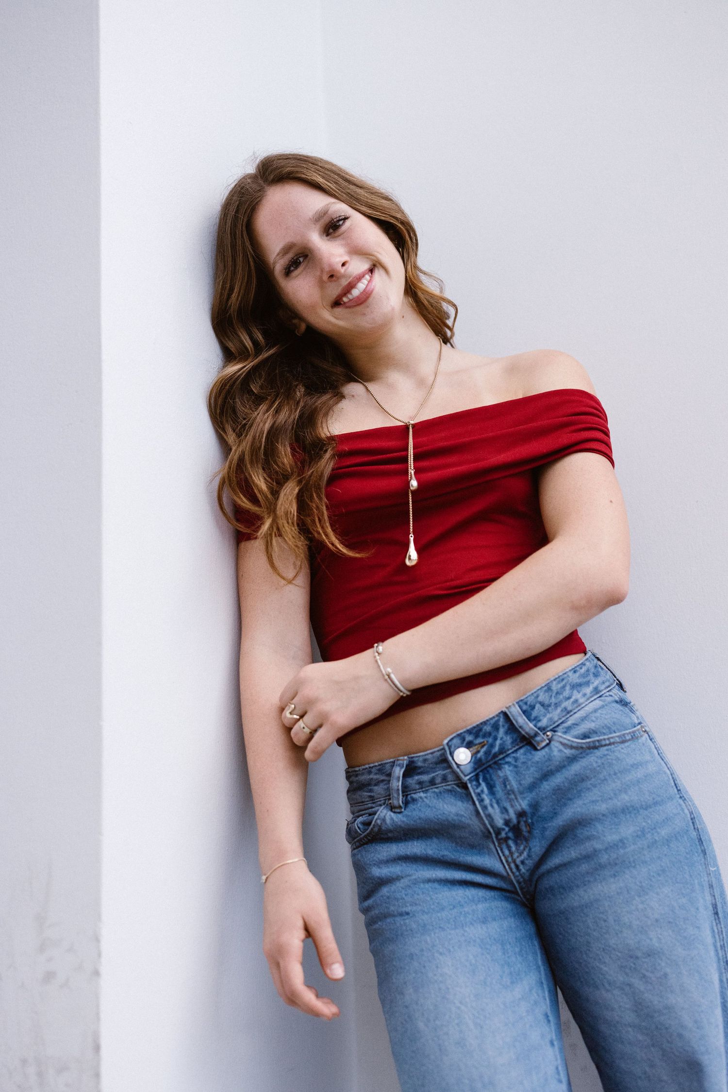Smiling woman in red off-shoulder top and jeans leaning against white wall.