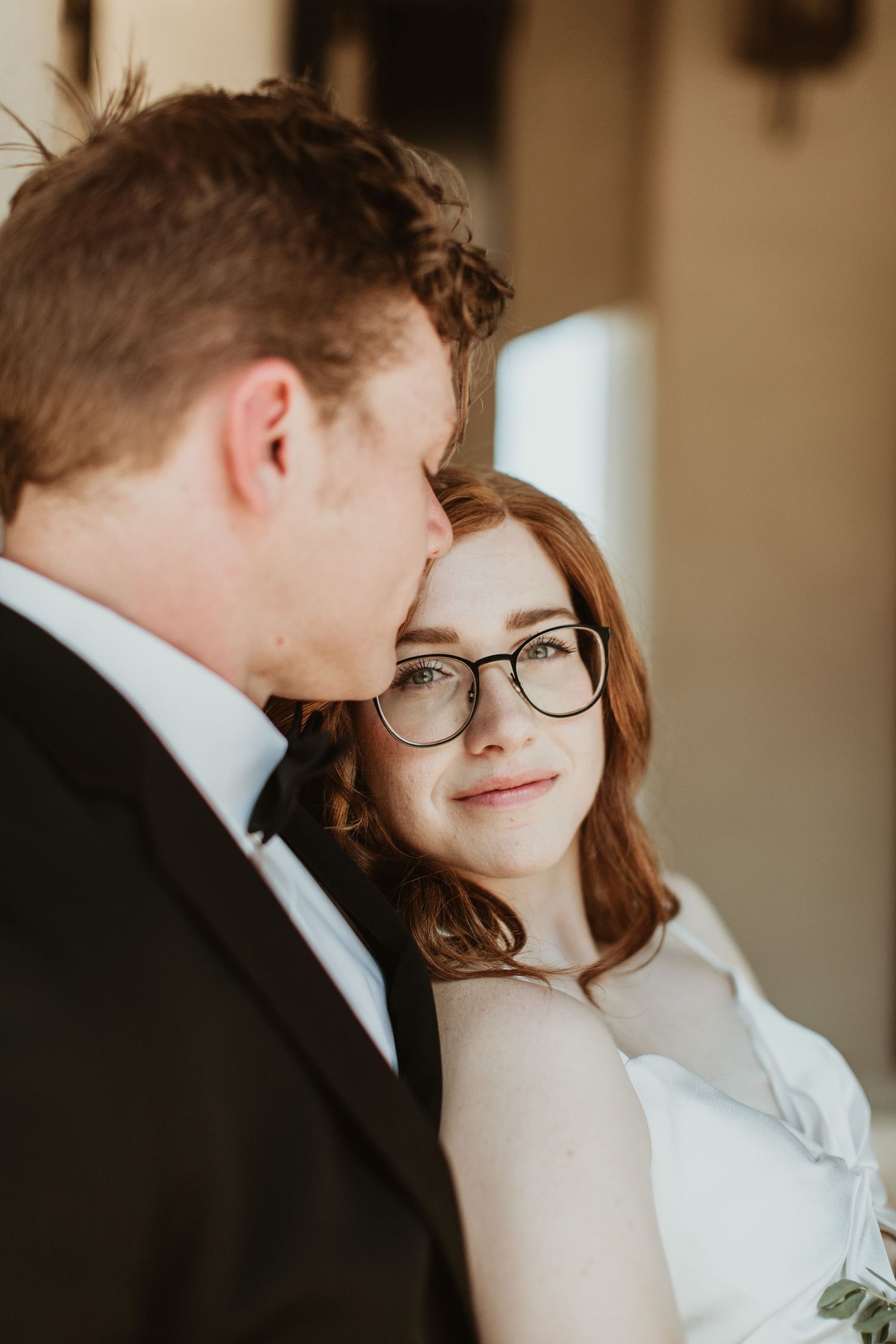 A bride with red hair and glasses smiles warmly while leaning against a groom in a black tuxedo.