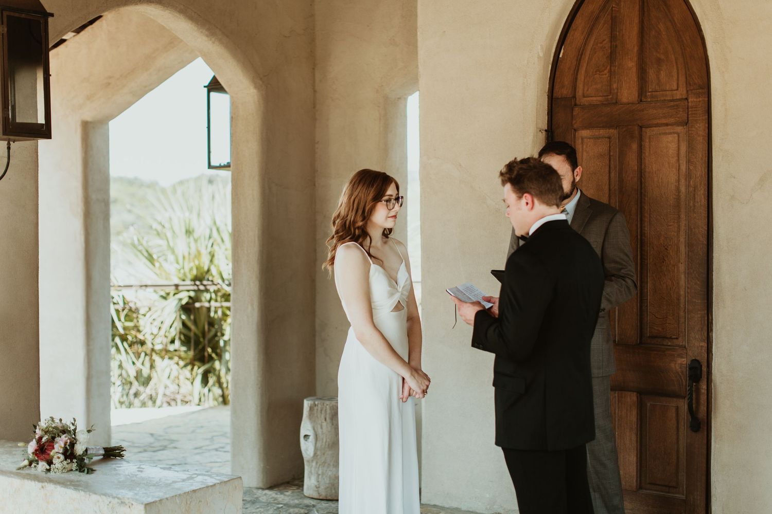 Couple exchanging wedding vows outdoors at a rustic venue with an arched doorway and wooden door.