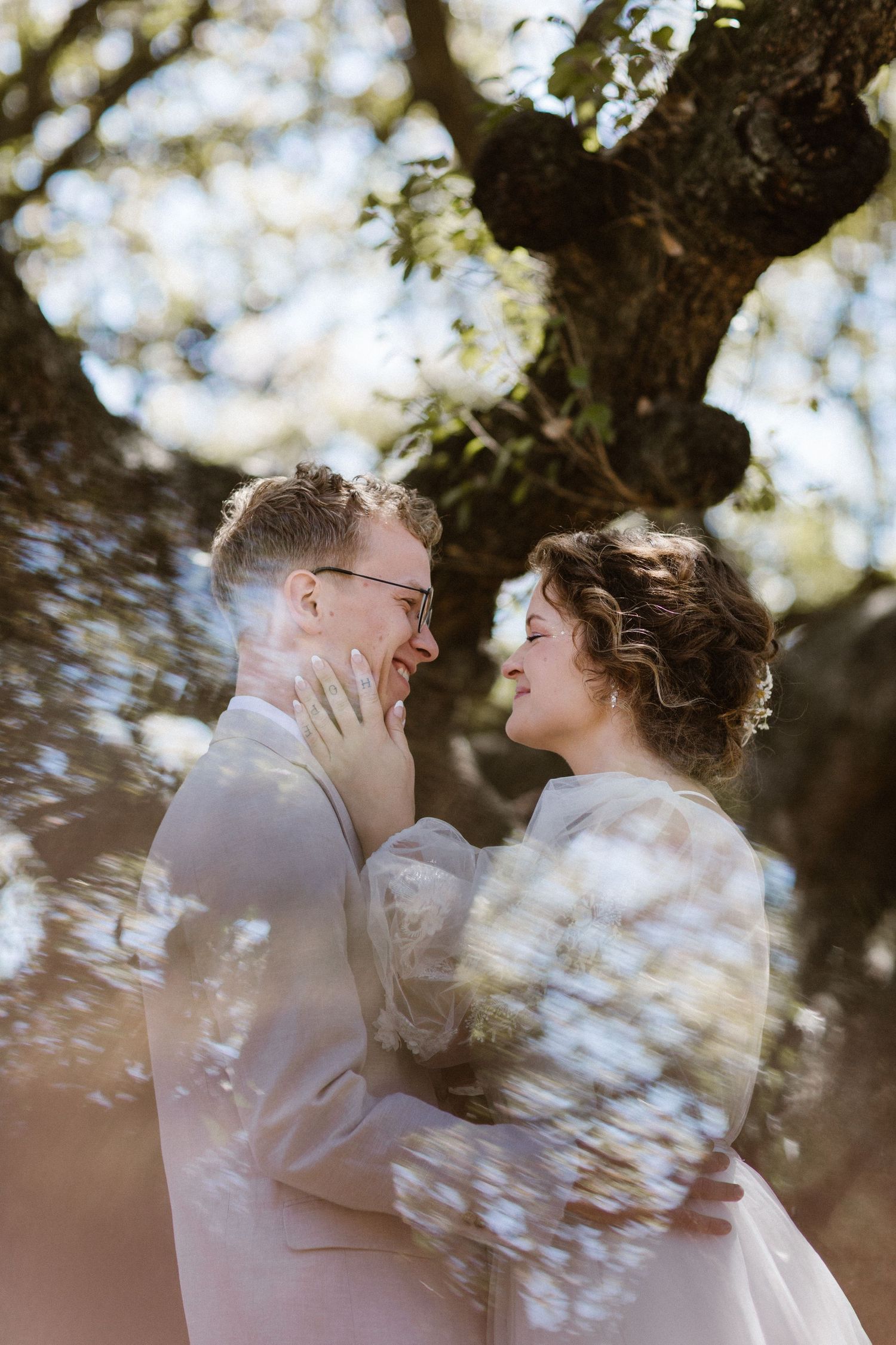 Bride and groom share a romantic moment under a sunlit oak tree, with soft bokeh floral effects framing their loving gaze.