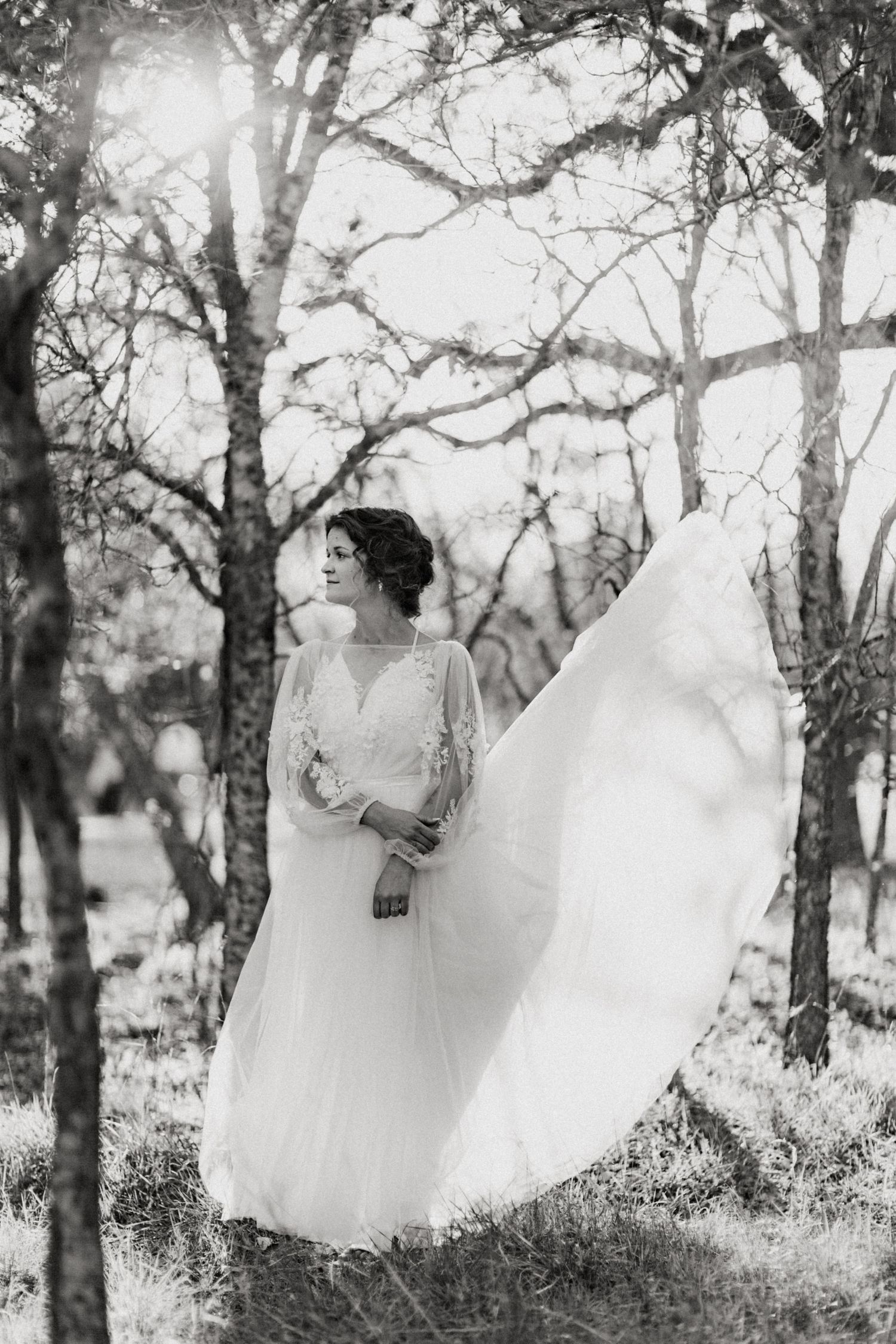 Black and white portrait of bride in flowing lace wedding dress standing among trees with veil blowing.