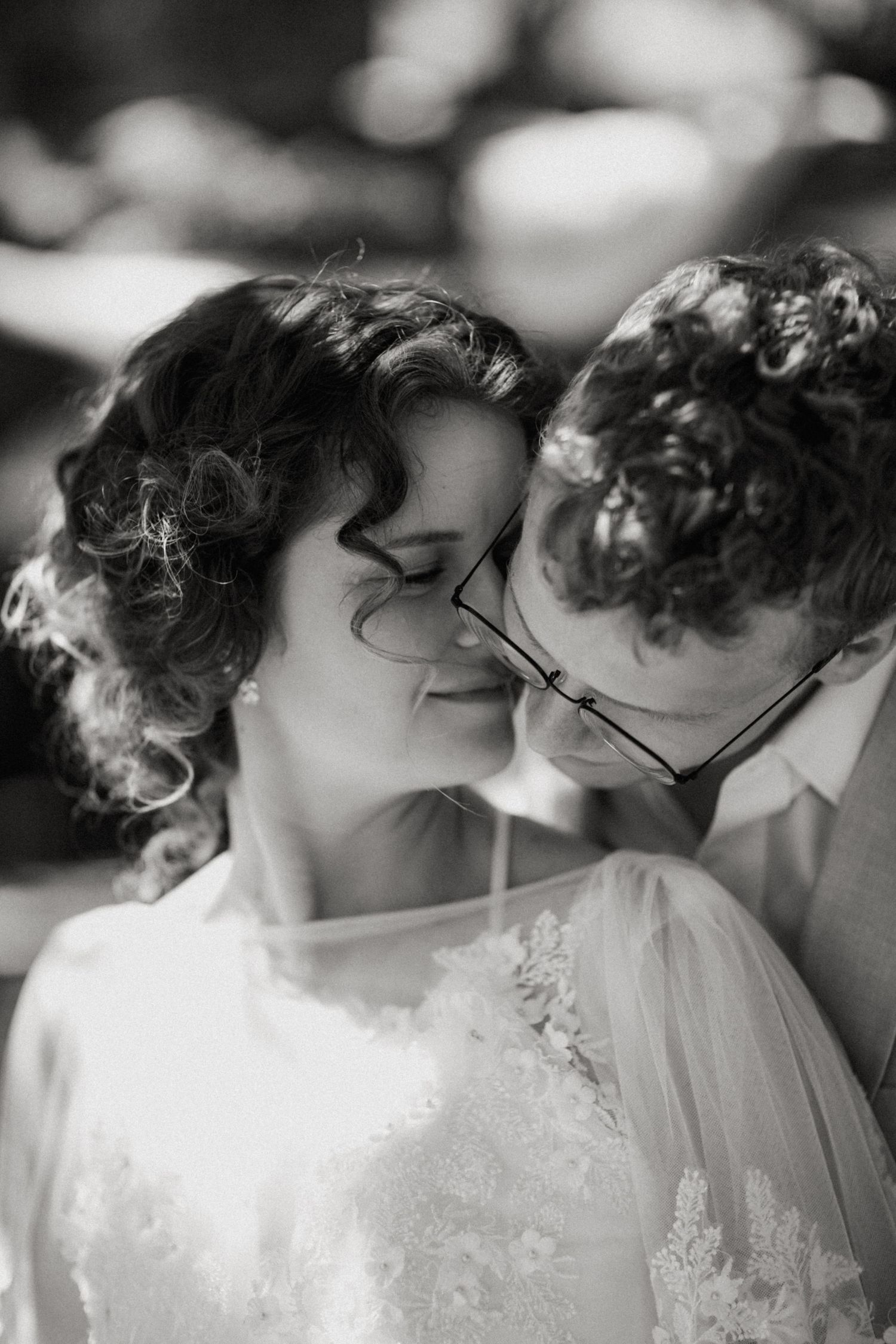Black and white wedding portrait of bride and groom sharing an intimate moment, groom wearing glasses nuzzling bride.