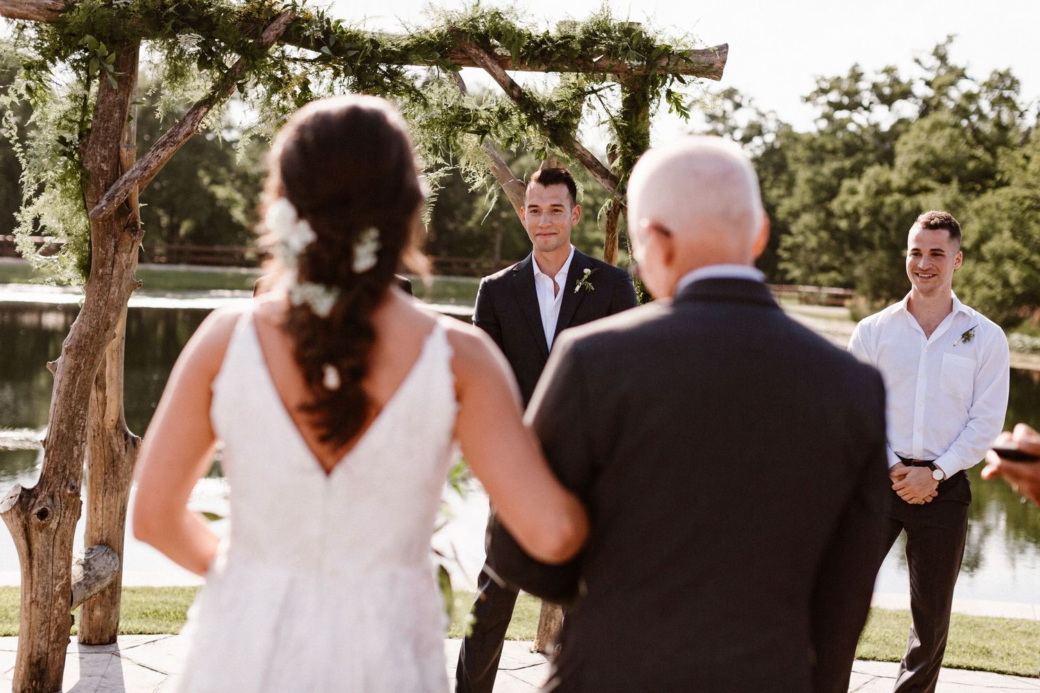 Groom smiling as bride walks down aisle at outdoor lakeside wedding ceremony.