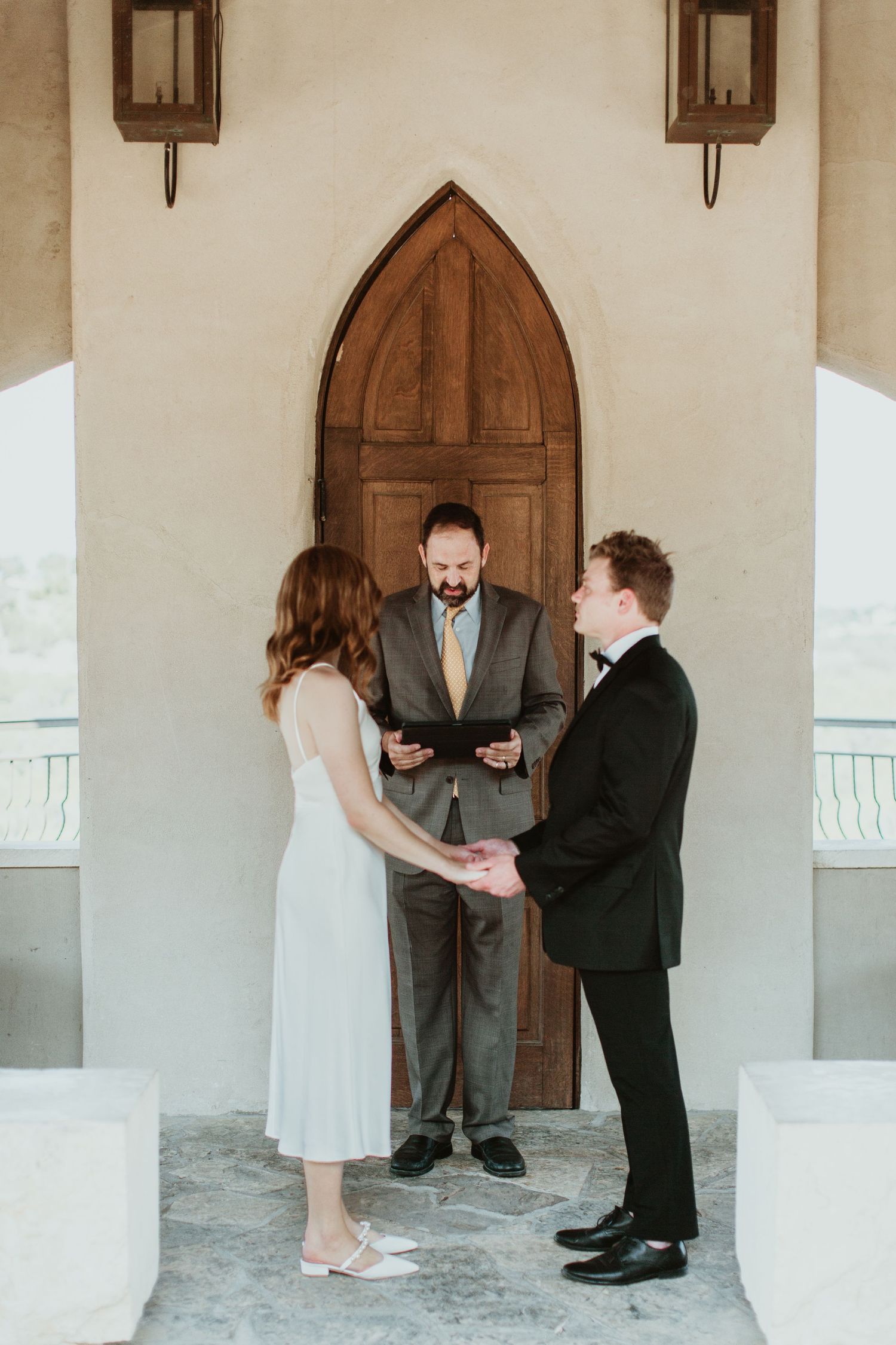 Couple exchanging vows at outdoor chapel ceremony with officiant holding book before arched wooden door.
