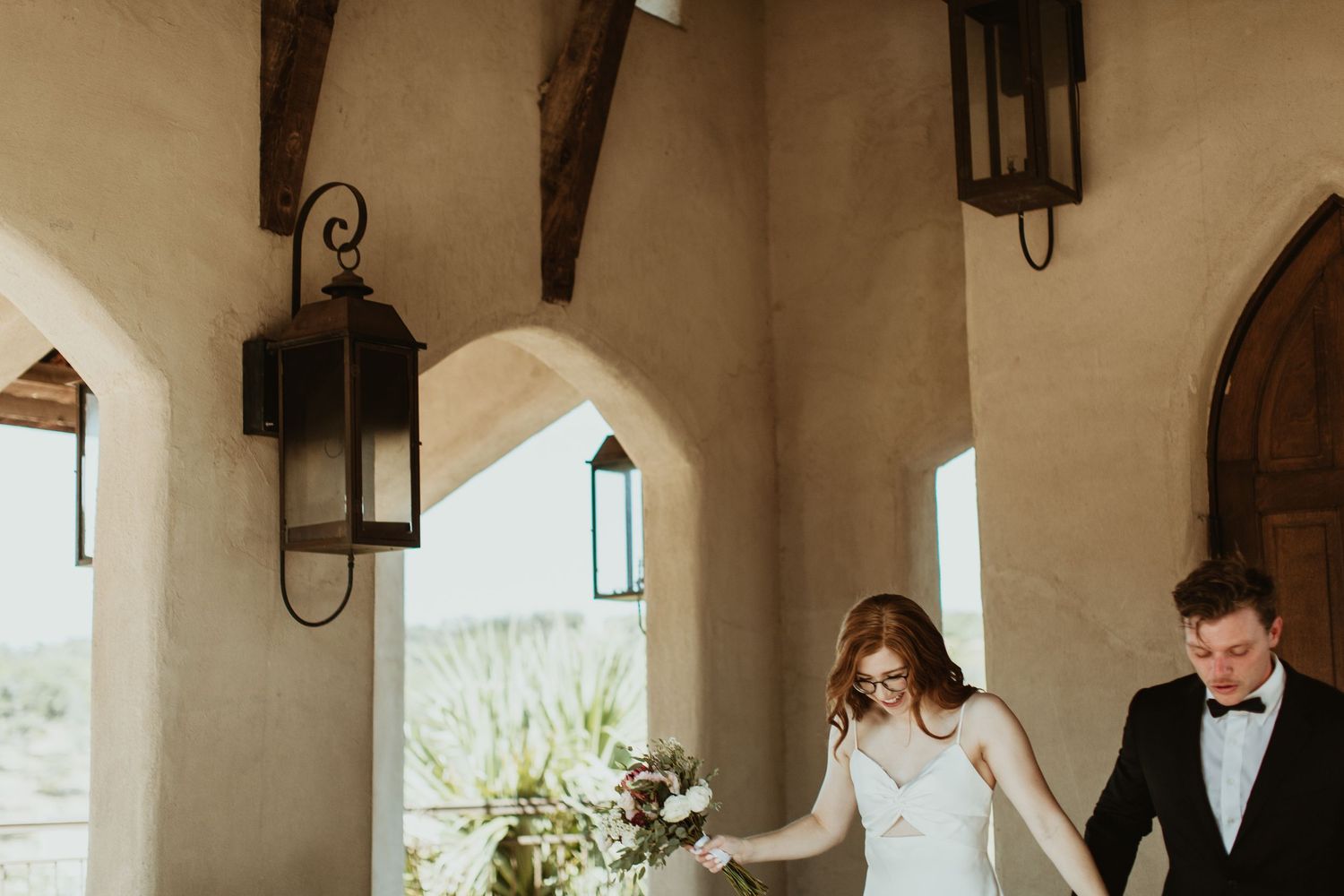 Bride and groom walk hand-in-hand under rustic arched portico with wrought iron lanterns at desert wedding venue.