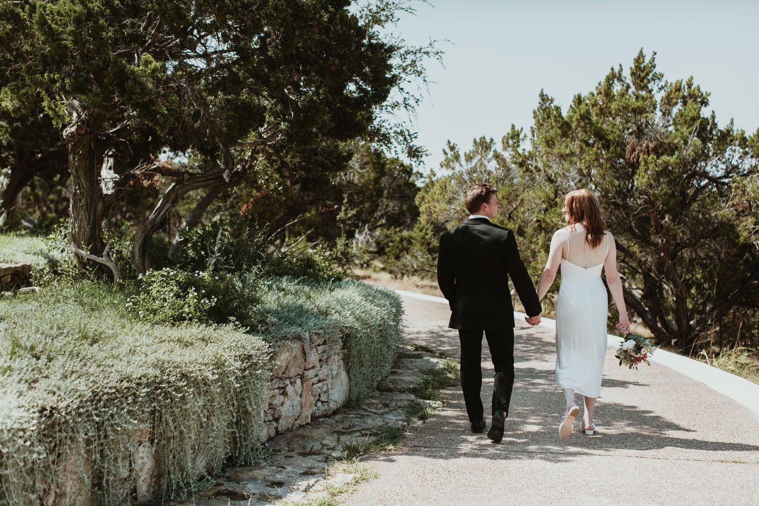 Bride and groom walking hand in hand down a scenic path lined with lush greenery and a stone wall on their wedding day.