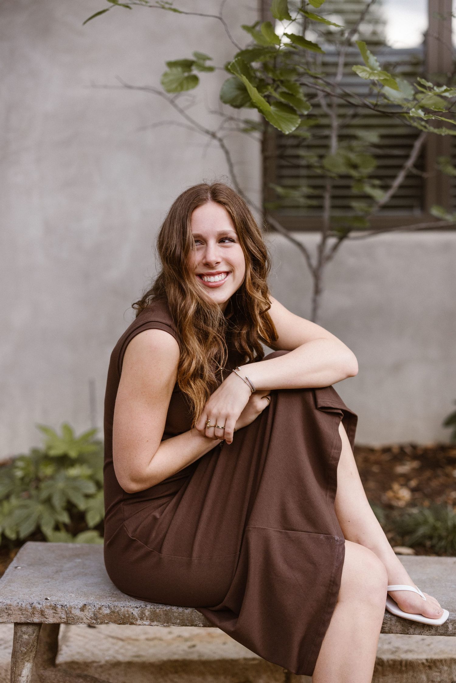 Smiling woman in brown sleeveless dress sitting on stone bench outdoors with greenery.