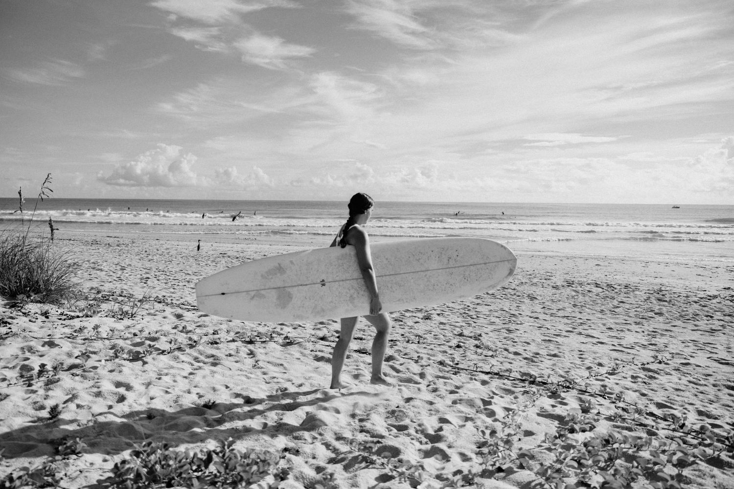Black and white photo of a woman carrying a longboard surfboard walking along a wide sandy beach.