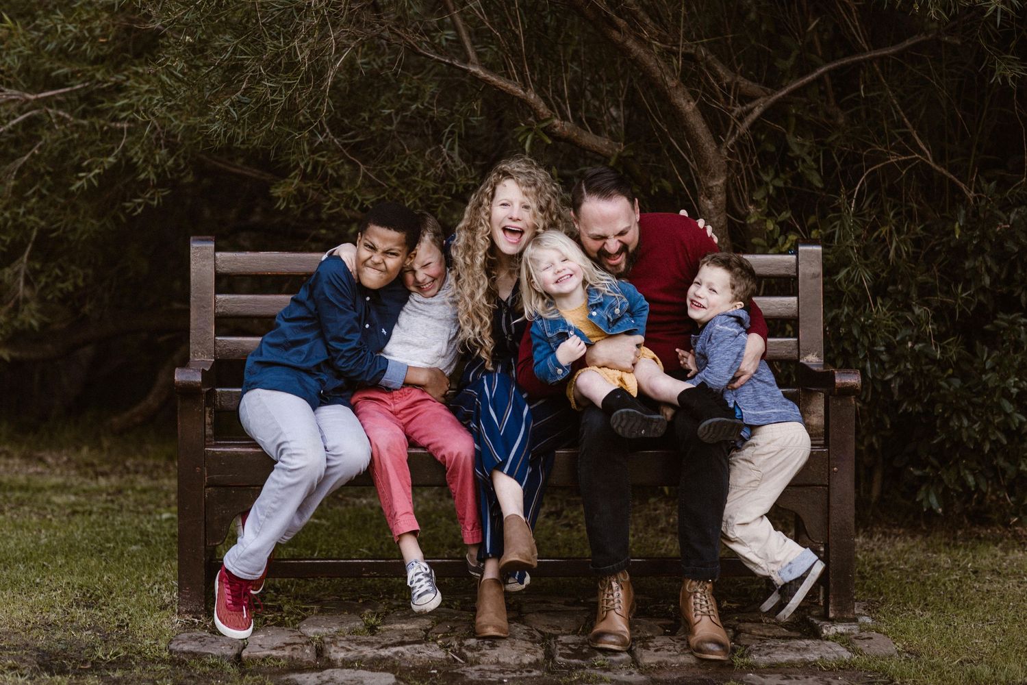 Joyful family of five laughing together on a wooden park bench surrounded by greenery.