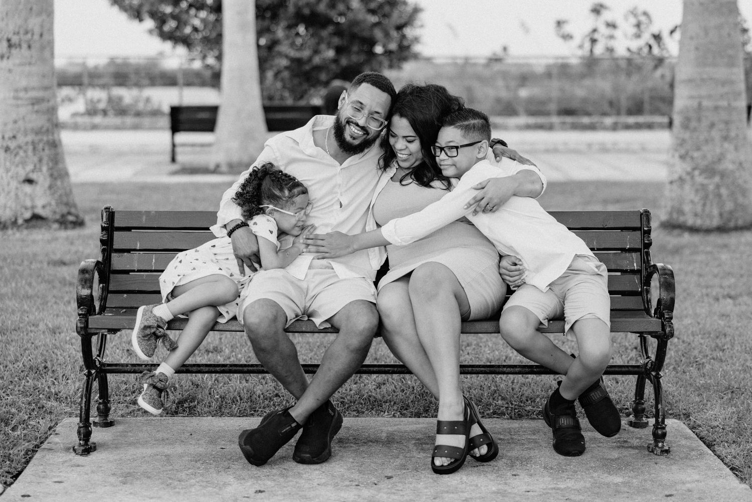 Black and white photo of a joyful family of four hugging on a park bench outdoors.