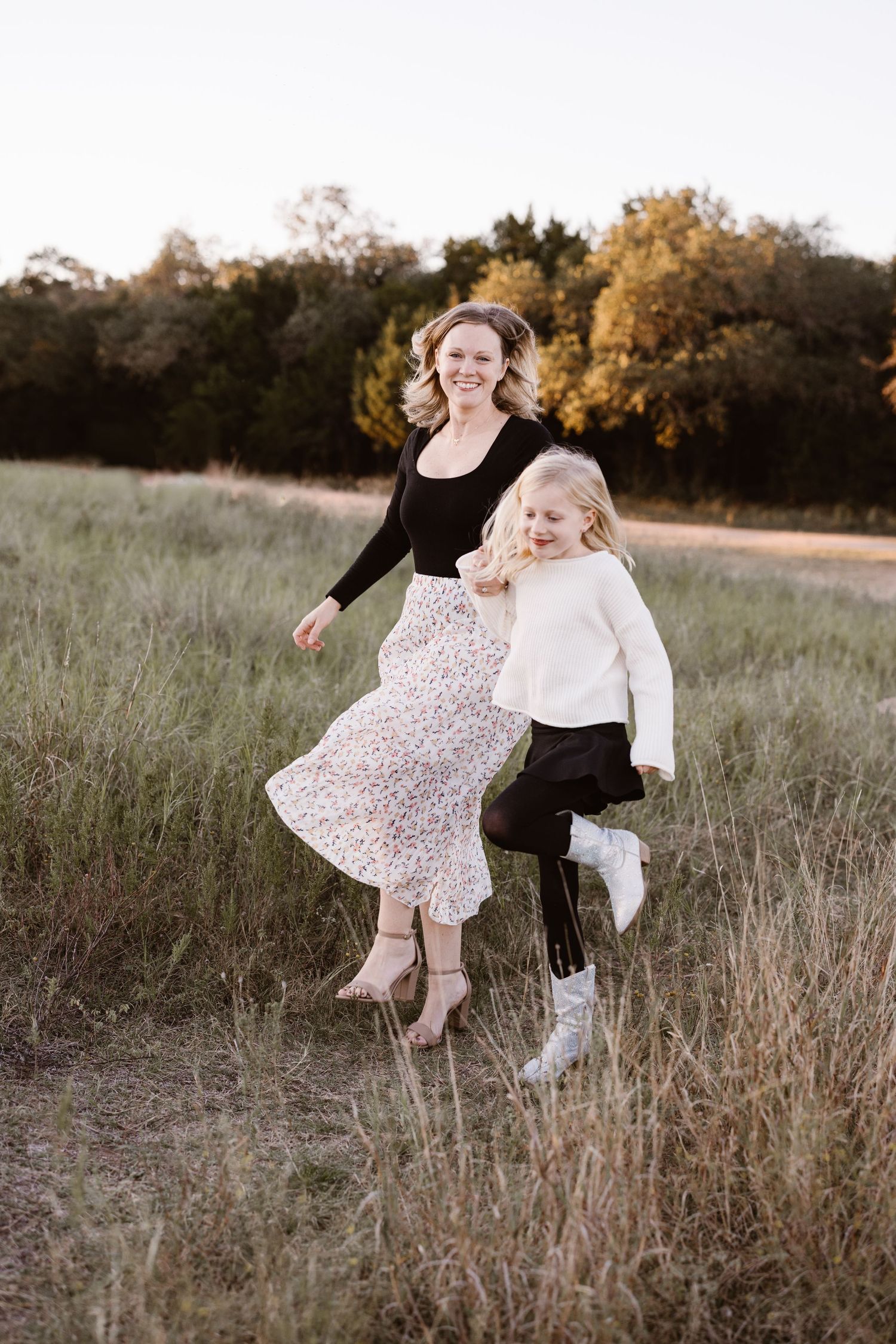Smiling mother and daughter skipping together through a grassy field at golden hour.