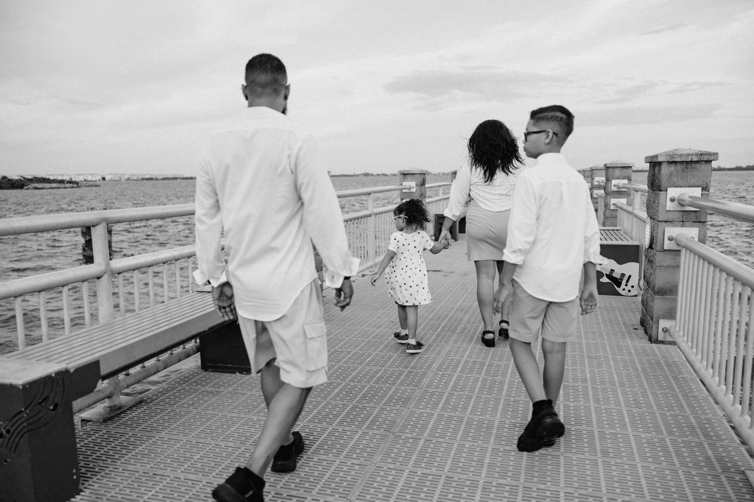 Black and white photo of family walking hand-in-hand along a wooden boardwalk by the water.