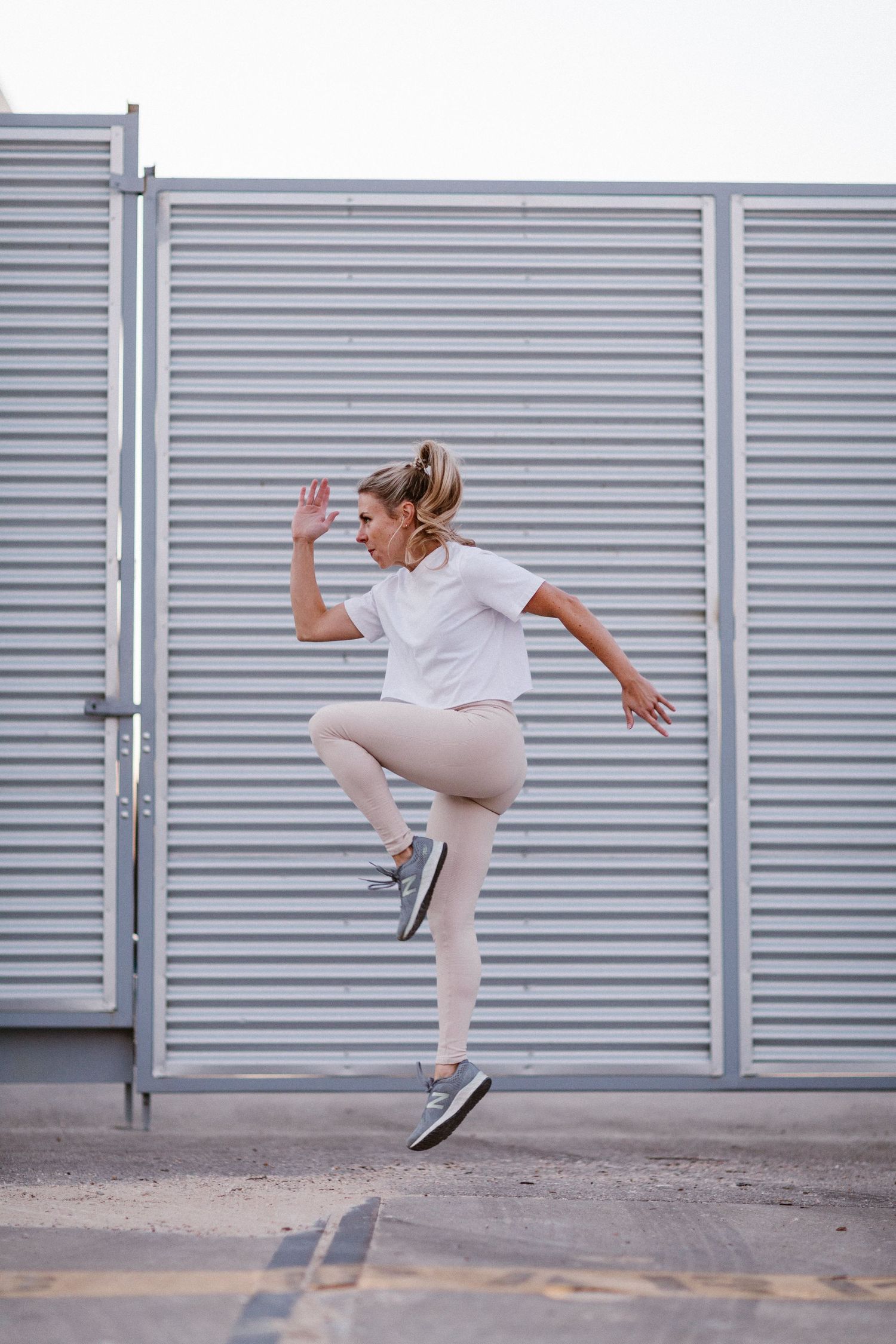 A woman in white athletic wear jumps dynamically in front of a corrugated metal wall outdoors.