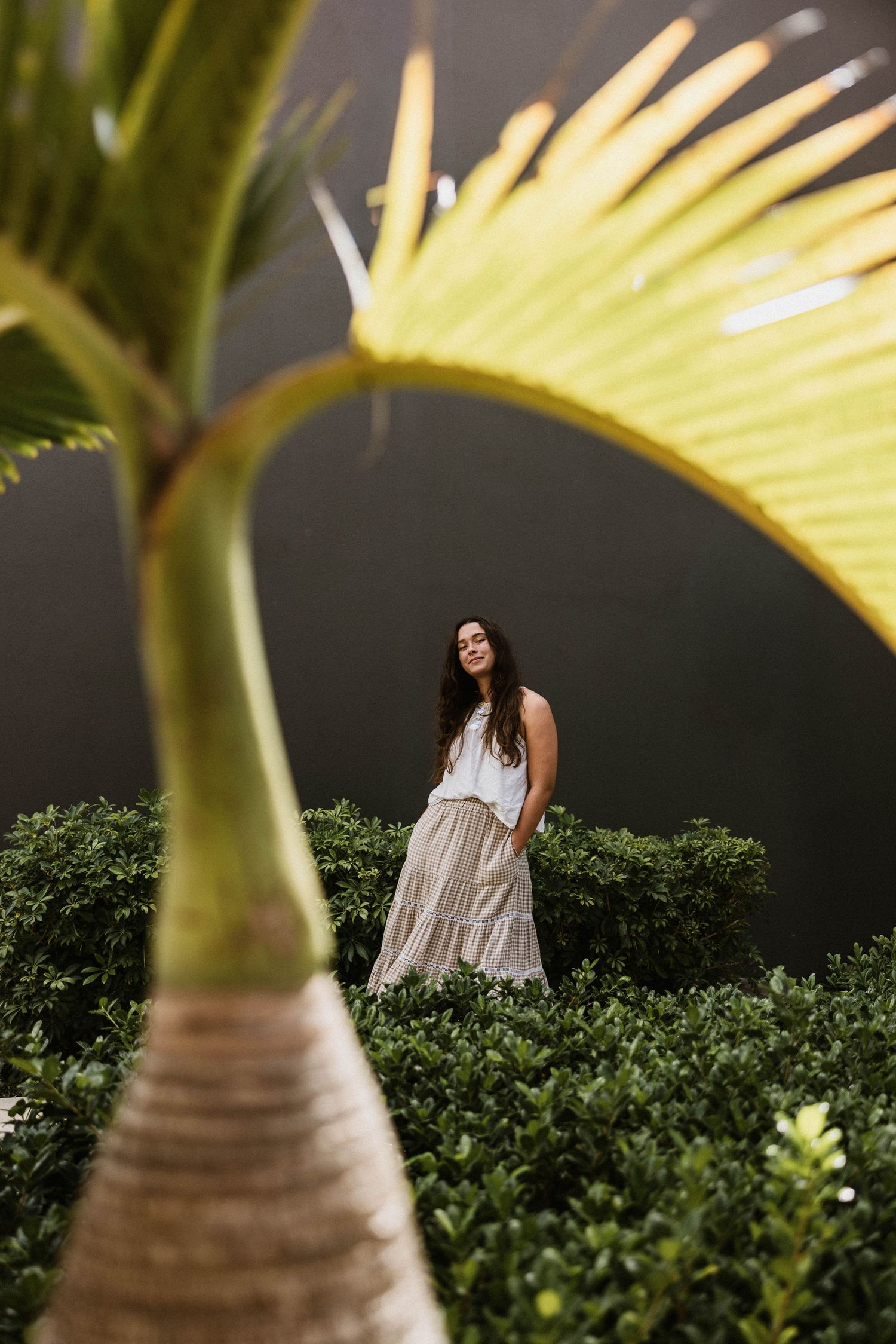 Young woman in a white top and tiered skirt standing among green hedges framed by a palm leaf.