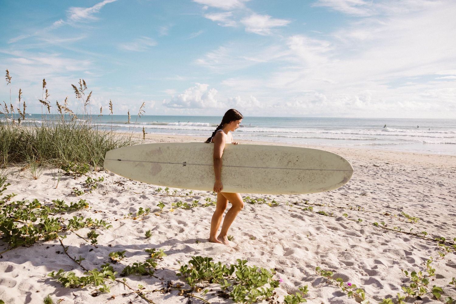 Woman in bikini carrying white surfboard walking toward ocean on sunny sandy beach.