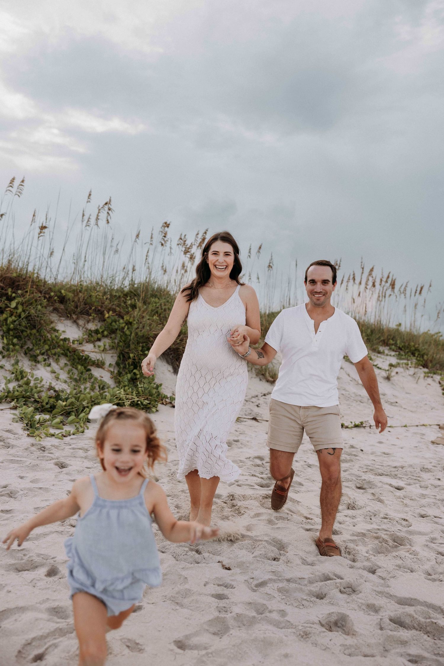 Joyful family running on a sandy beach, with a pregnant mother in a white dress and playful toddler.