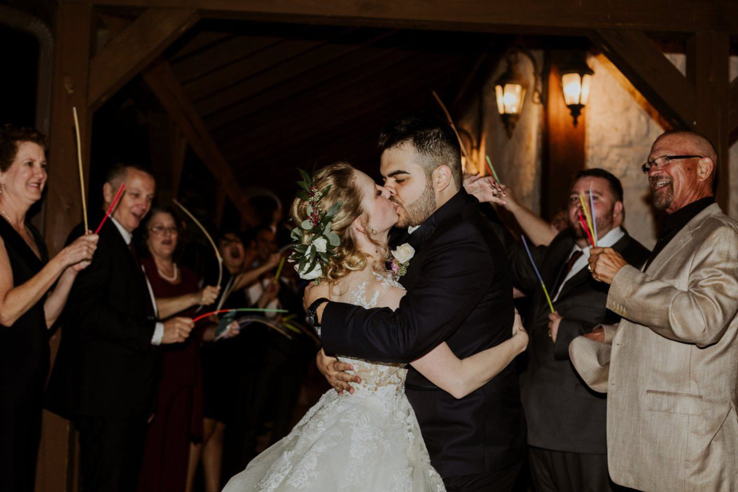 Bride and groom share a romantic kiss during their wedding sparkler send-off surrounded by cheering guests.