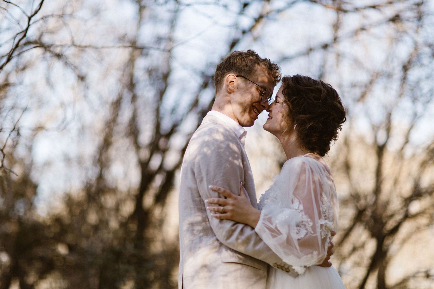 Bride and groom embracing closely and touching foreheads beneath bare winter trees in soft light.