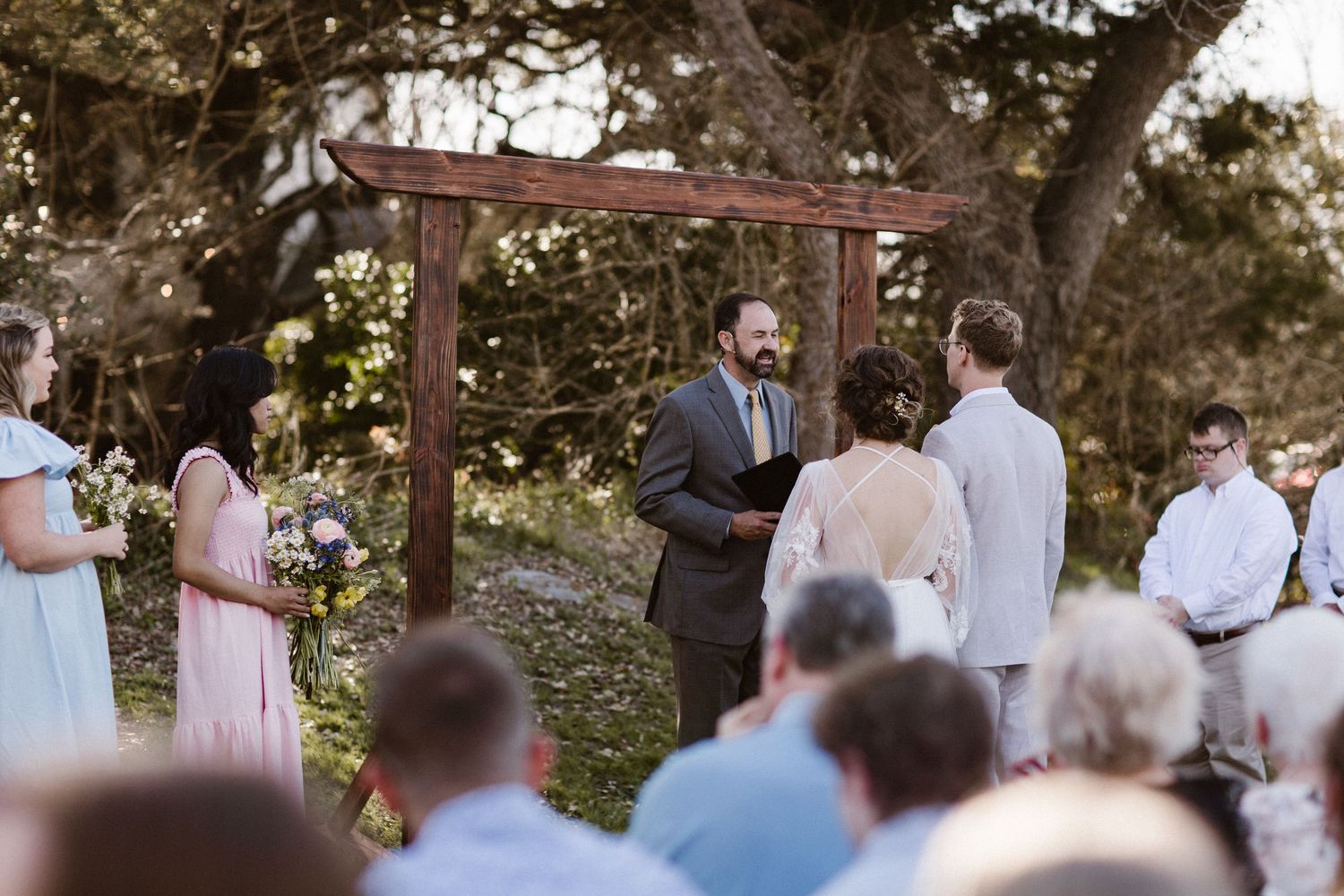 Outdoor wedding ceremony under rustic wooden arch with officiant, bride, groom, and guests gathered among trees.