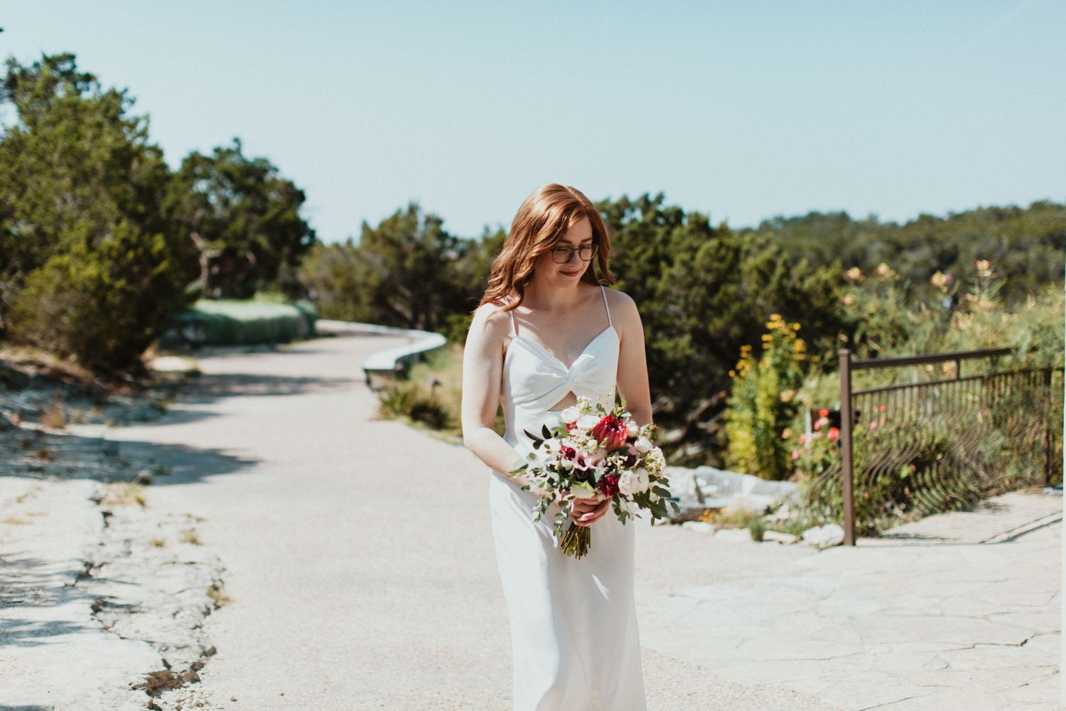 Bride in white gown holding pink floral bouquet walks sandy path surrounded by lush greenery on sunny wedding day.
