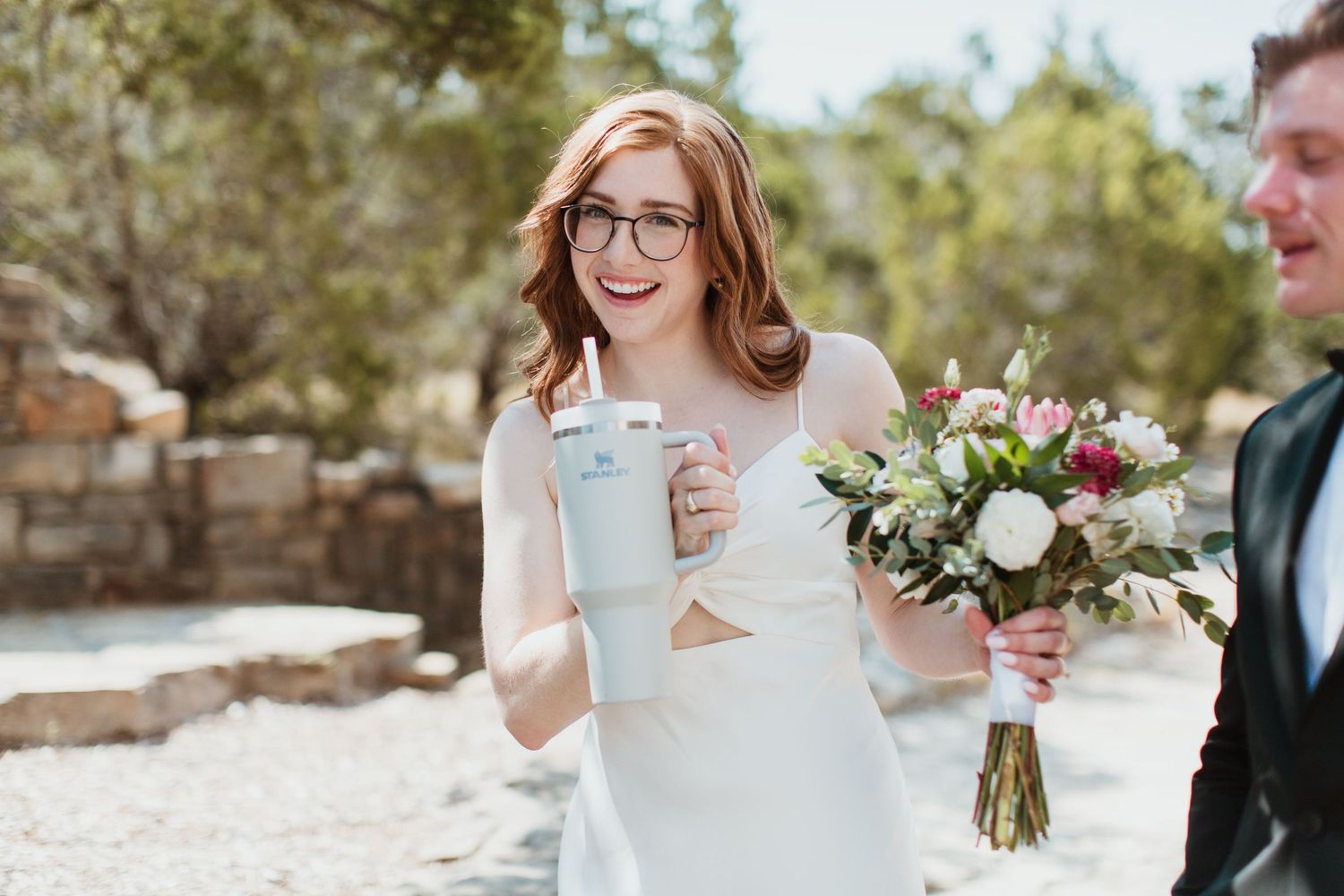 Smiling bride in white dress holds bouquet and Stanley tumbler cup at outdoor rustic wedding celebration.