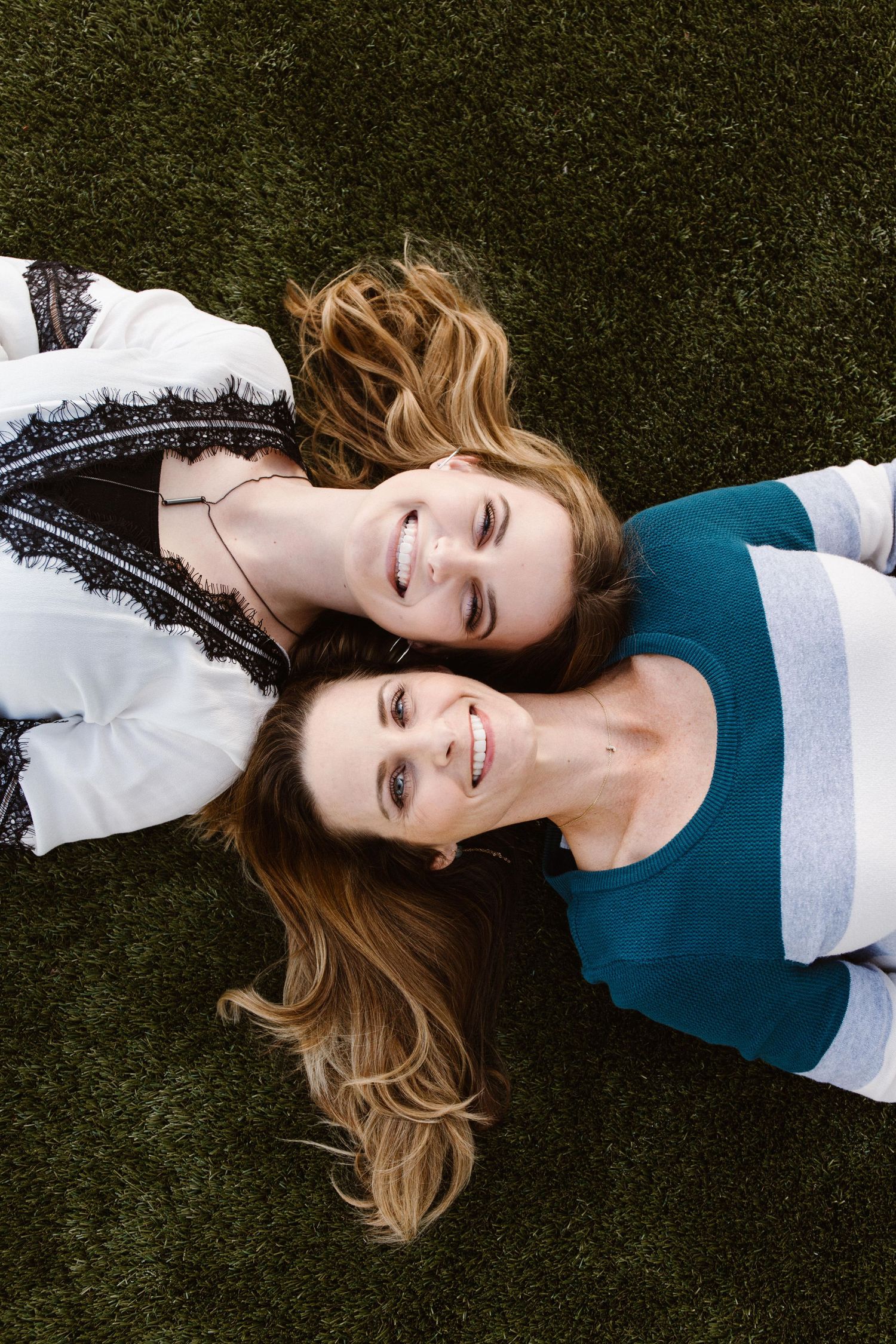Two smiling young women lying head-to-head on green grass looking up at camera.