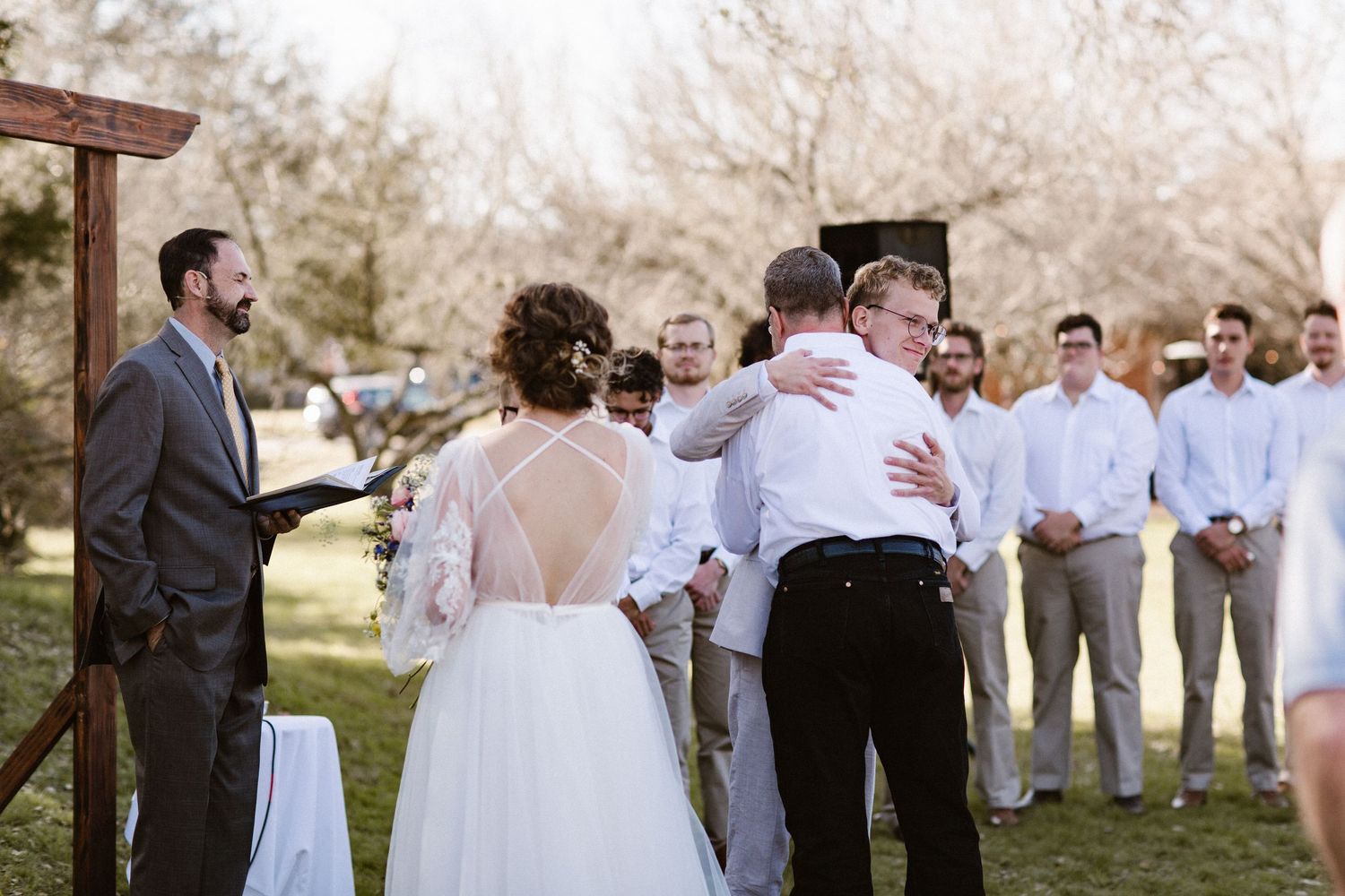 Bride and groom share a warm hug with a guest at the altar following their outdoor wedding ceremony under a wooden arch.