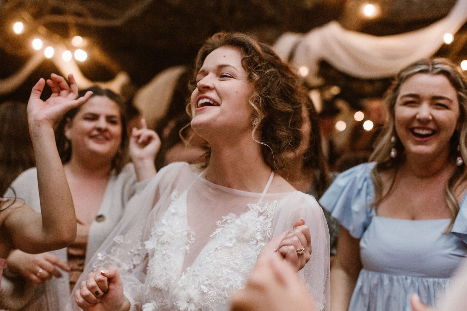 Bride in white lace gown laughing joyfully while dancing with guests at wedding reception.