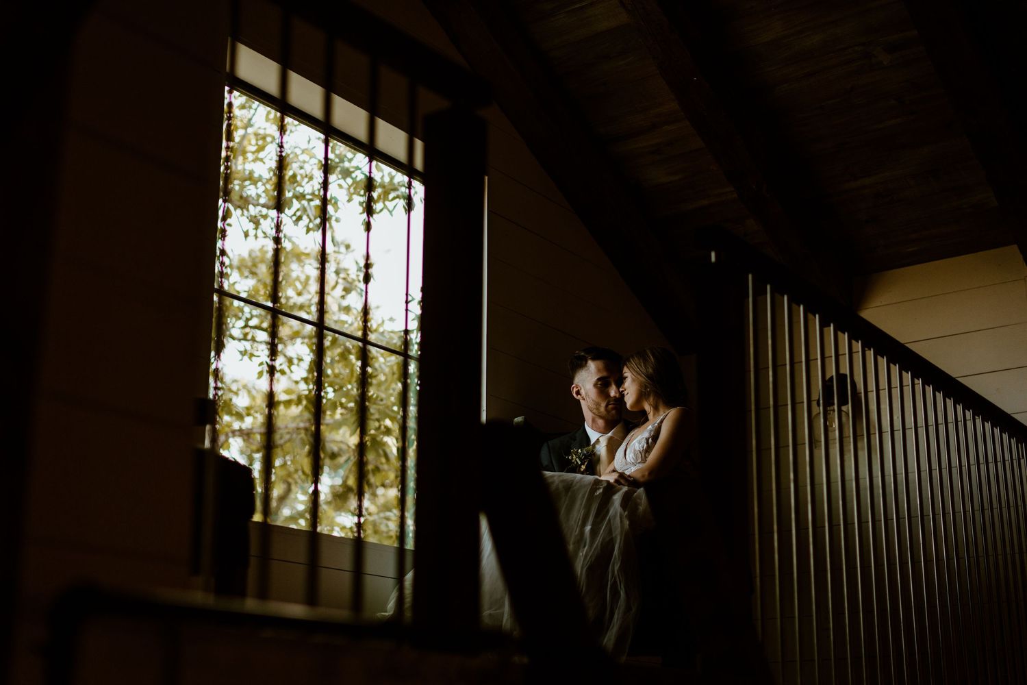 Bride and groom share an intimate moment by a large window in a dimly lit rustic venue.