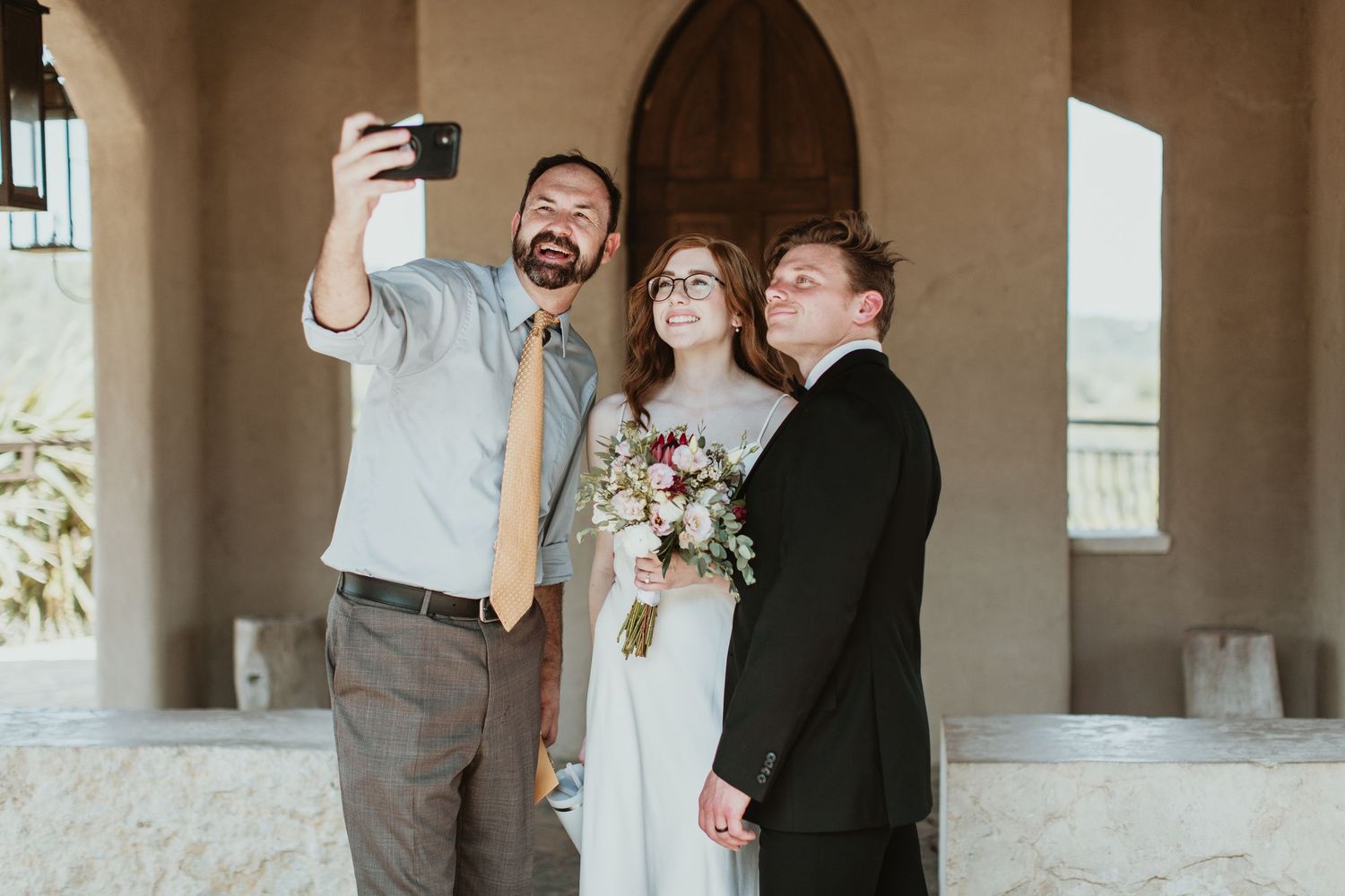 A groom takes a selfie with a bride holding a bouquet and a guest in formal attire at a wedding venue.