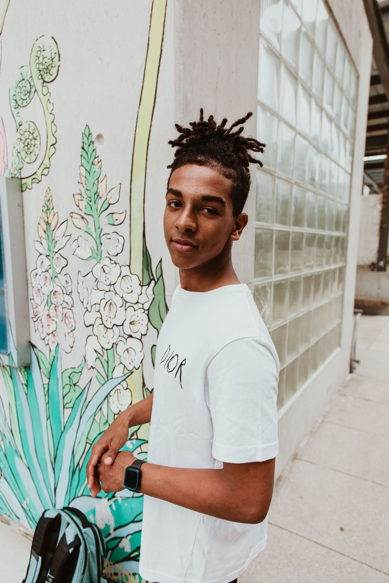 Young man with dreadlocks wearing a white Dior t-shirt standing beside a colorful floral mural outdoors.
