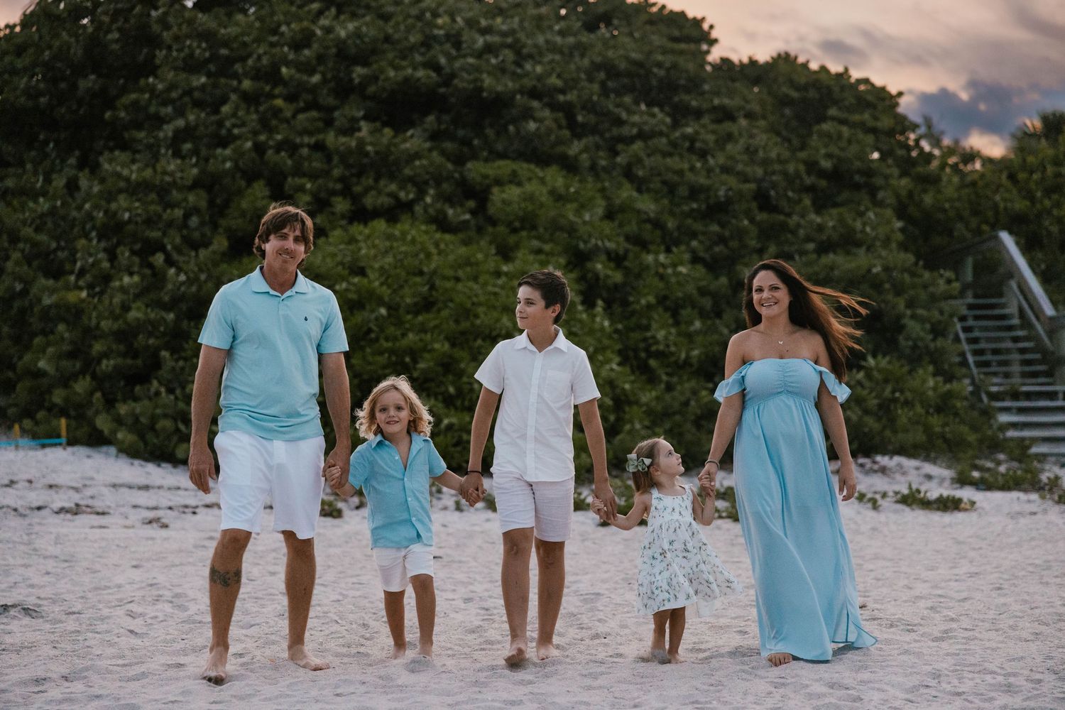 Family of five in coordinating blue and white outfits walking on sandy beach.