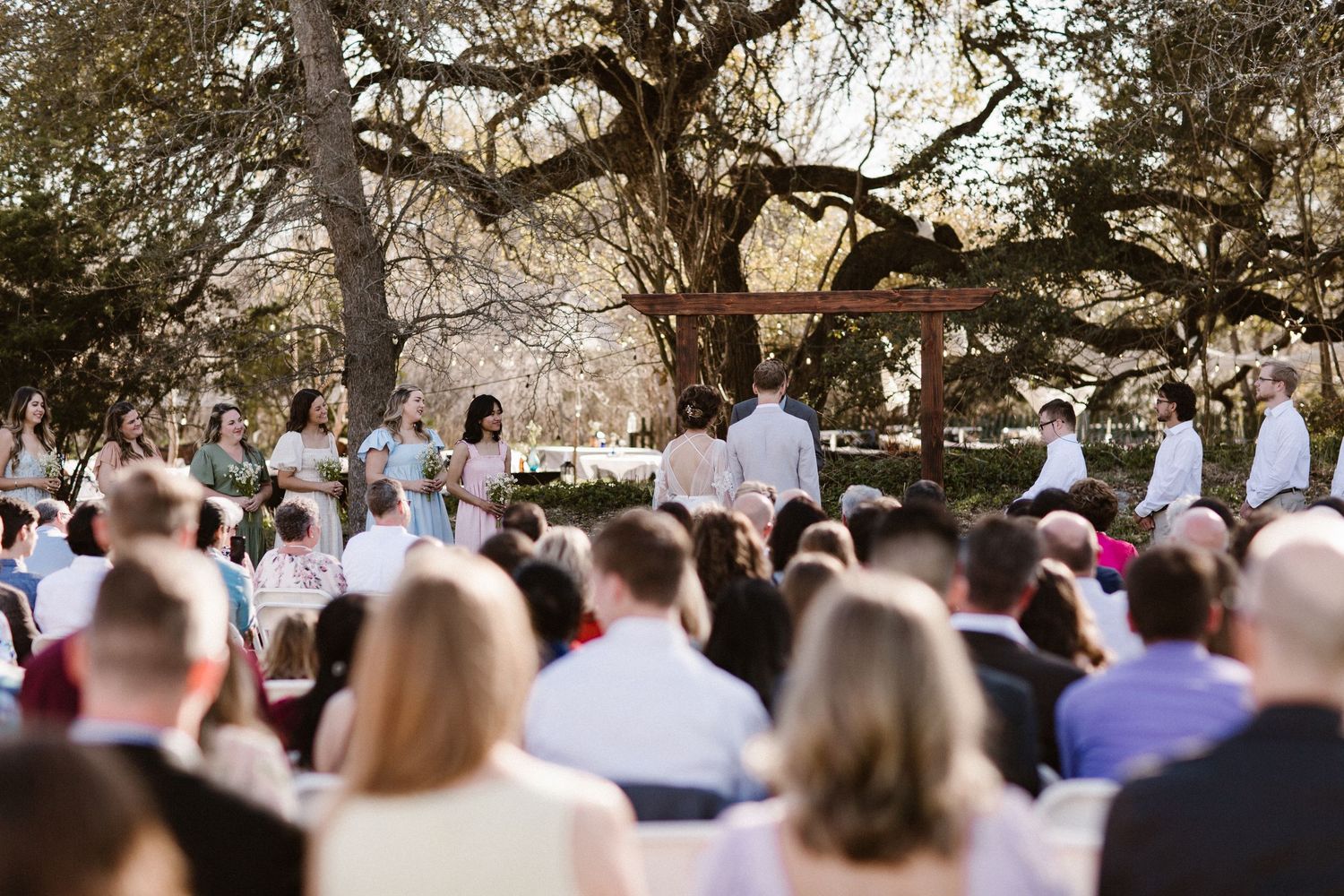 Wedding party stands at the altar during an outdoor ceremony beneath sprawling oak trees.