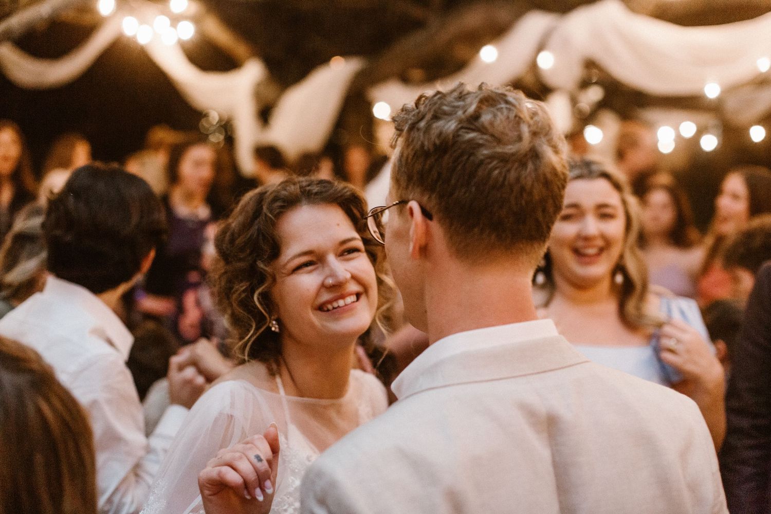 Bride smiling joyfully at groom during wedding reception dance under string lights.