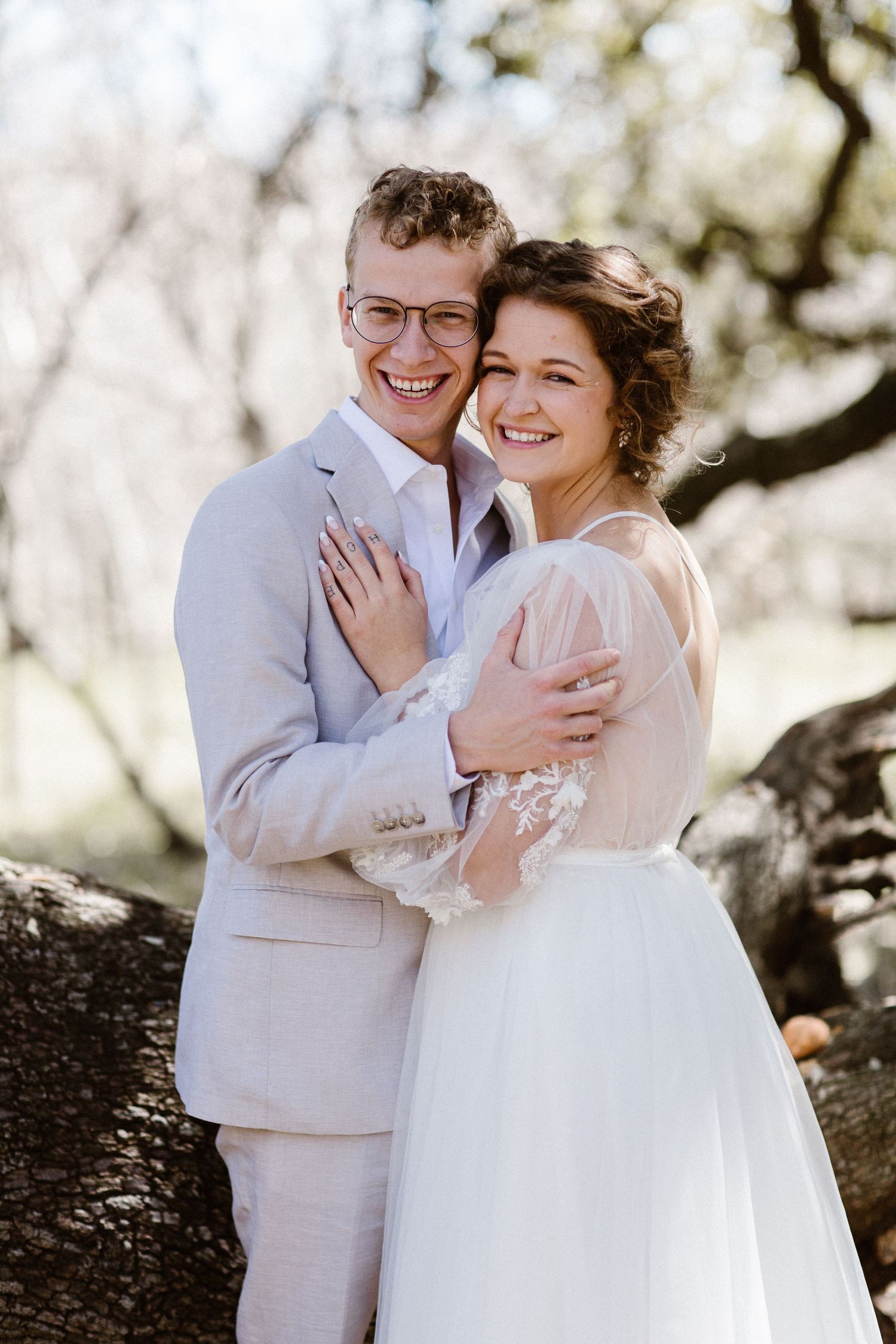 Smiling bride and groom share joyful embrace in sunlit outdoor wedding portrait among trees.