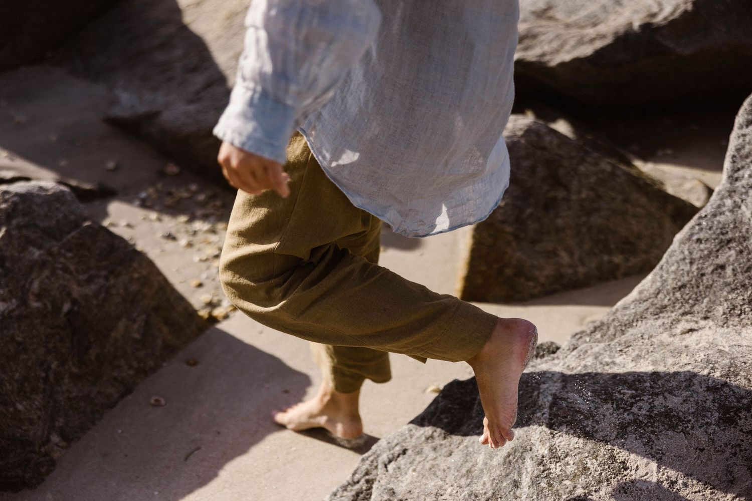 Barefoot person walking on rocks wearing olive linen pants and white linen shirt at the beach.