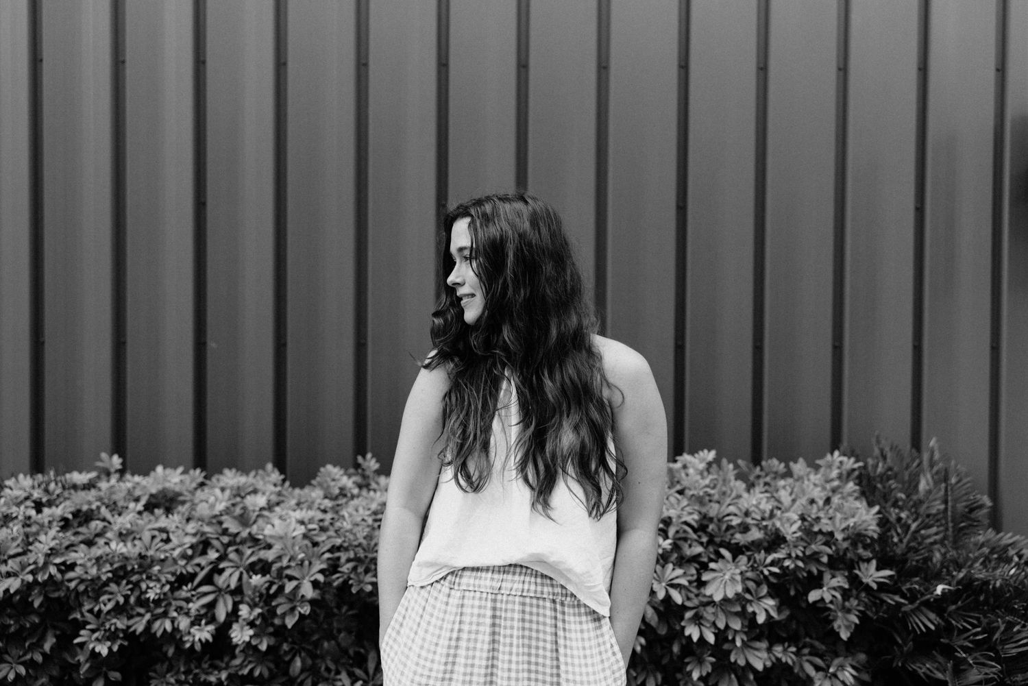 Young woman with long dark hair smiling outdoors in black and white, wearing a white top and plaid skirt.