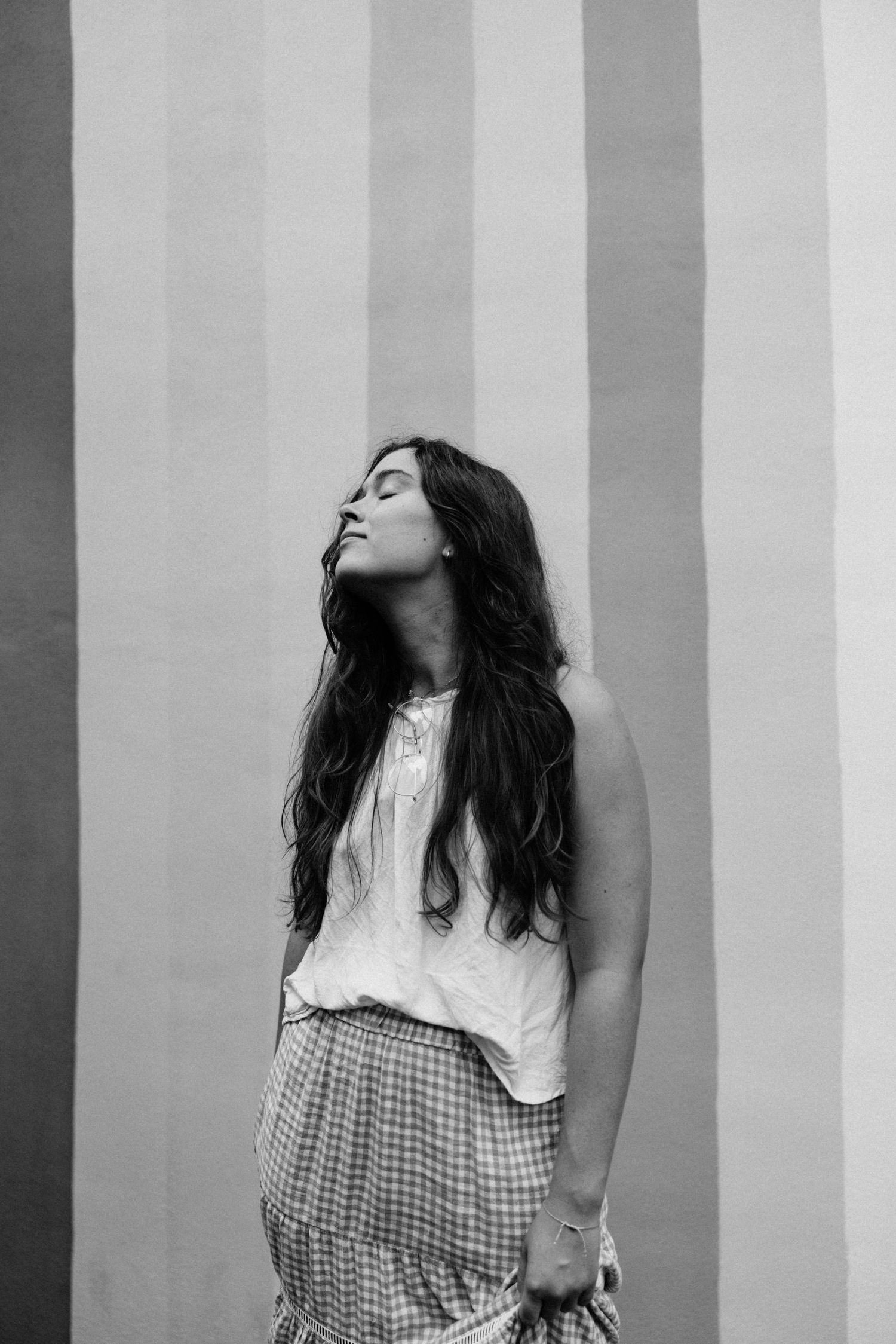 Black and white portrait of a young woman with long hair standing against a striped wall.