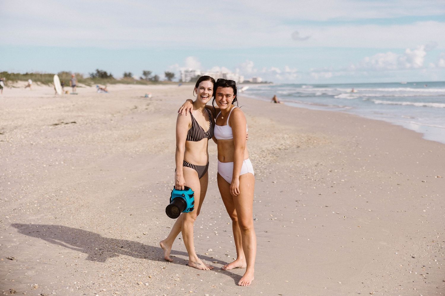 Two women in bikinis pose together on a sunny sandy beach holding a camera.