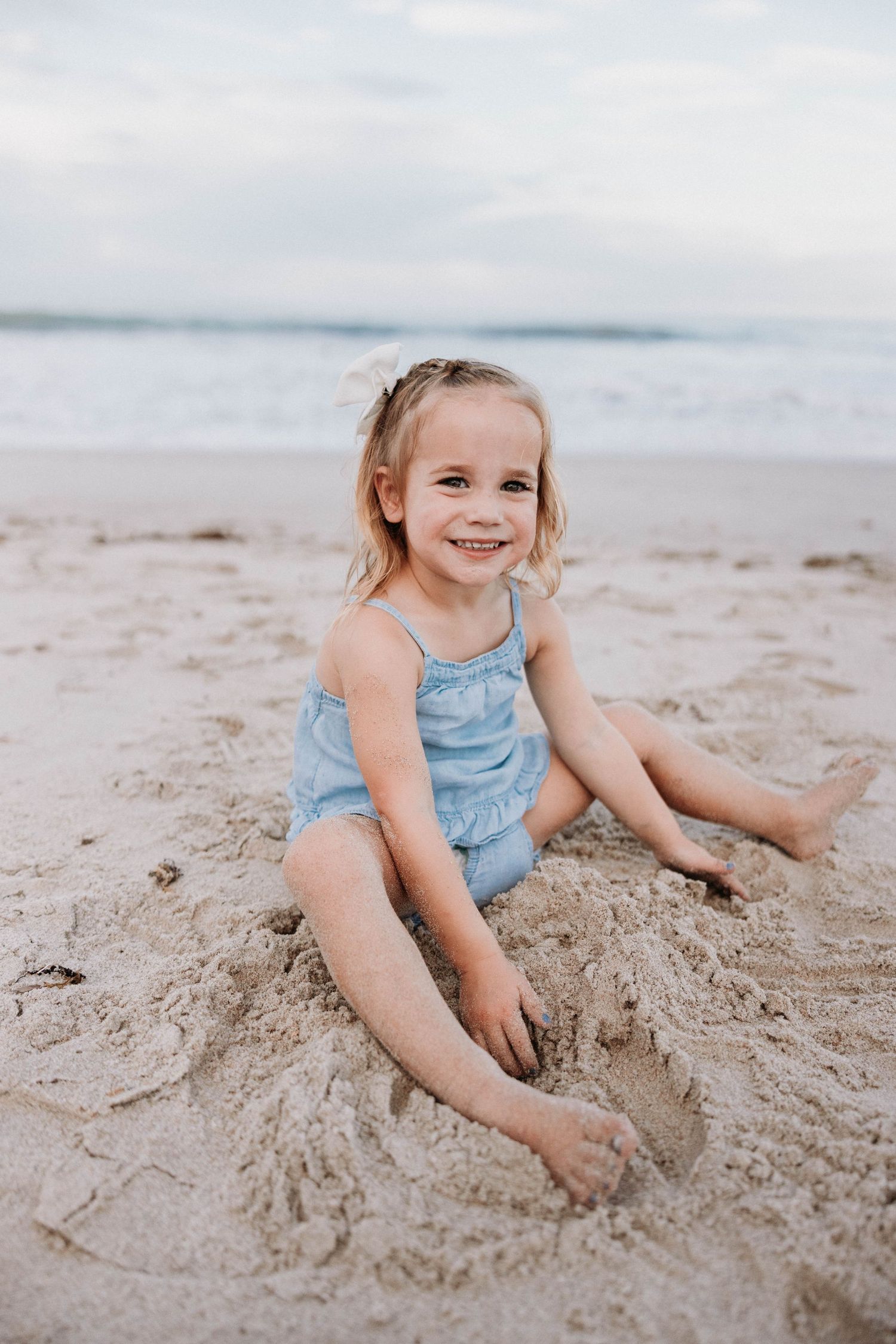 Adorable smiling toddler girl in blue swimsuit sitting in the sand at the beach.