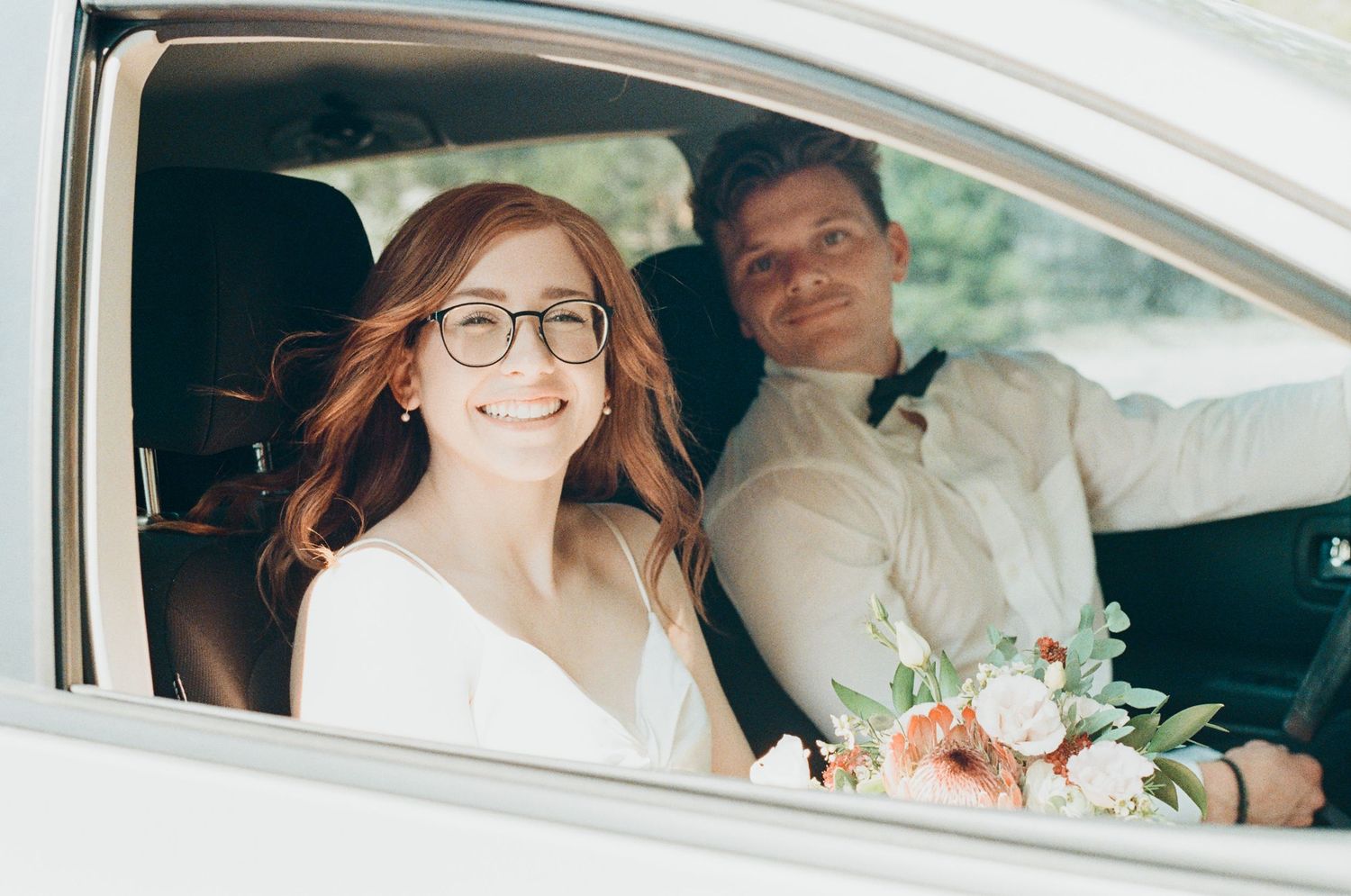 Smiling bride with glasses holding a floral bouquet sits in a white car beside a groom in a white shirt and bow tie.