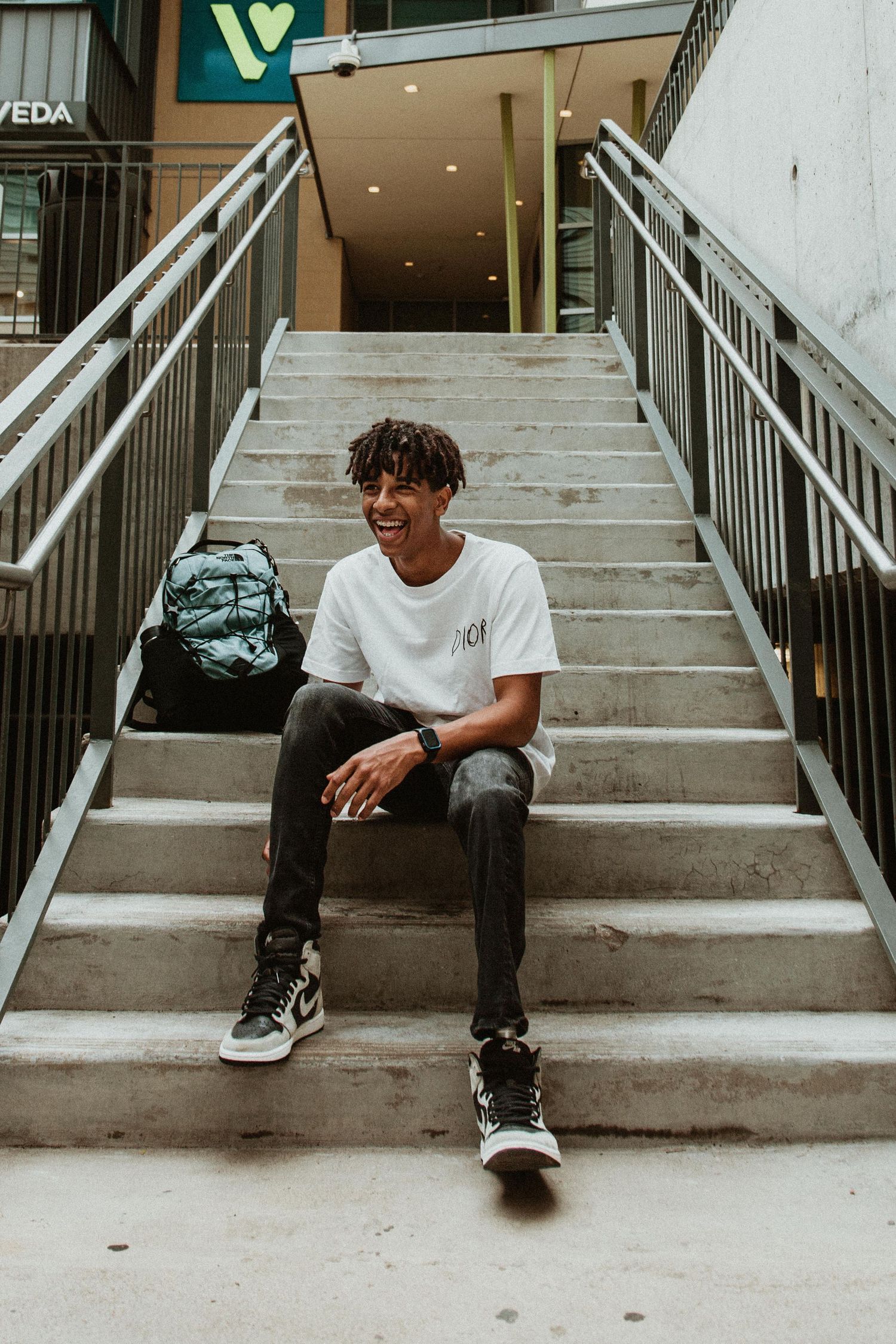 Young man smiling on concrete steps outdoors, wearing white t-shirt, dark jeans, and high-top sneakers, with backpack beside him.